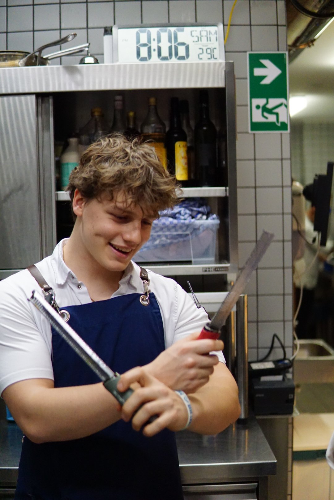 A young man with curly hair wearing a white shirt and apron, holding two kitchen tools, smiling in a commercial kitchen with shelves, bottles, and a digital clock showing 8:06 AM and 29°C in the background.