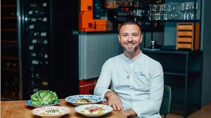 A man in a white chef's coat sitting at a table with various dishes, in a modern kitchen or restaurant setting.