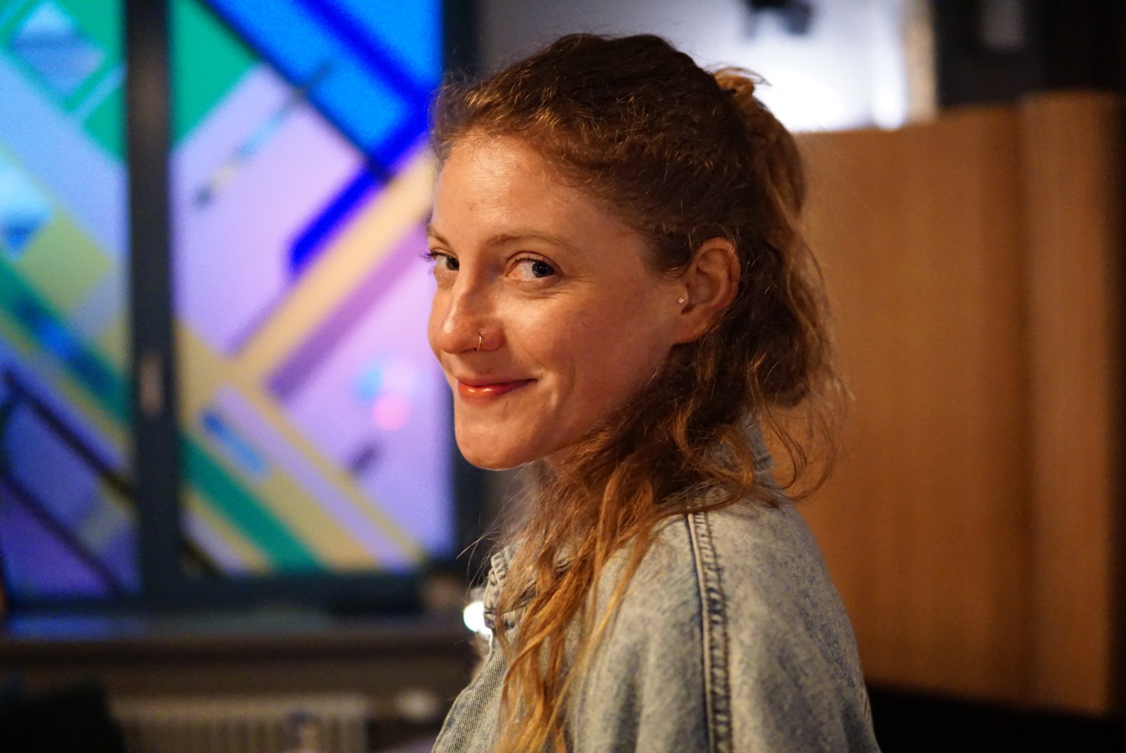 A woman with curly reddish-brown hair, wearing a denim jacket, smiling and looking over her shoulder in a dimly lit room with colorful geometric wall art.