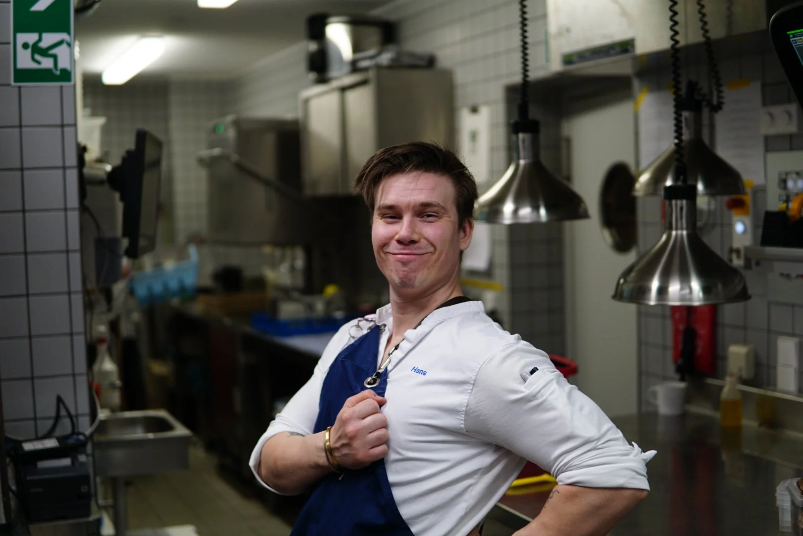 A chef smiling confidently in a commercial kitchen, wearing a white chef's coat and a blue apron, with stainless steel kitchen equipment and hanging lamps in the background.