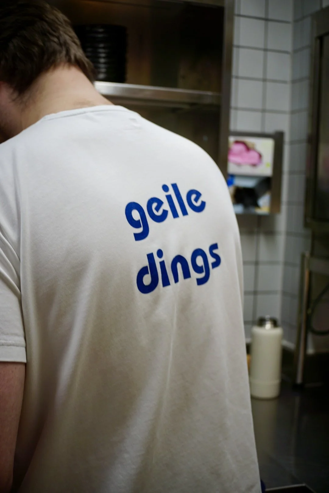 Back view of a person wearing a white T-shirt with blue text reading 'geile dinges' in a kitchen or restaurant setting.