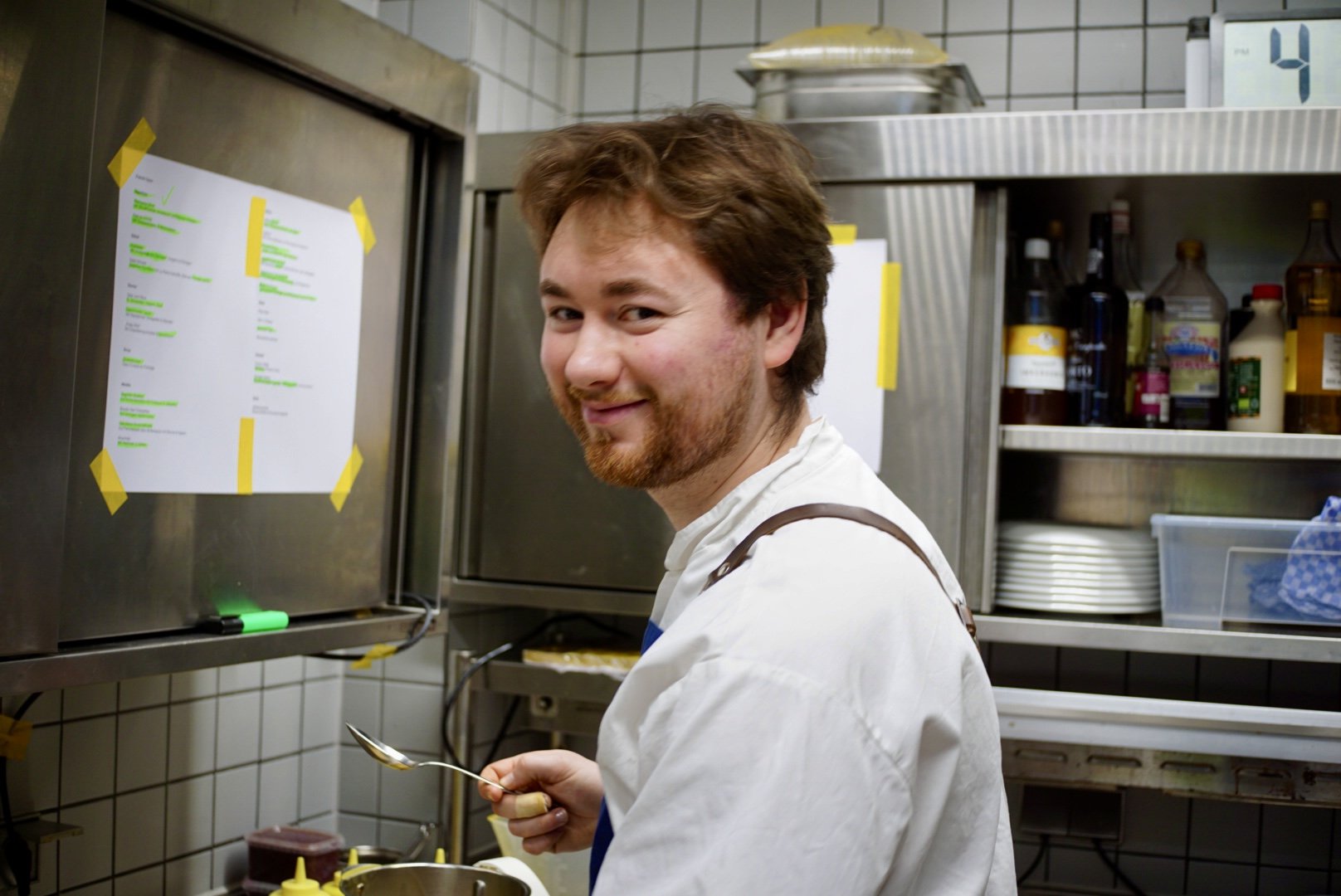 A man with brown hair and a beard, wearing a white shirt, stands in a commercial kitchen holding a spoon and smiling at the camera, with kitchen shelves and a menu on the wall behind him.