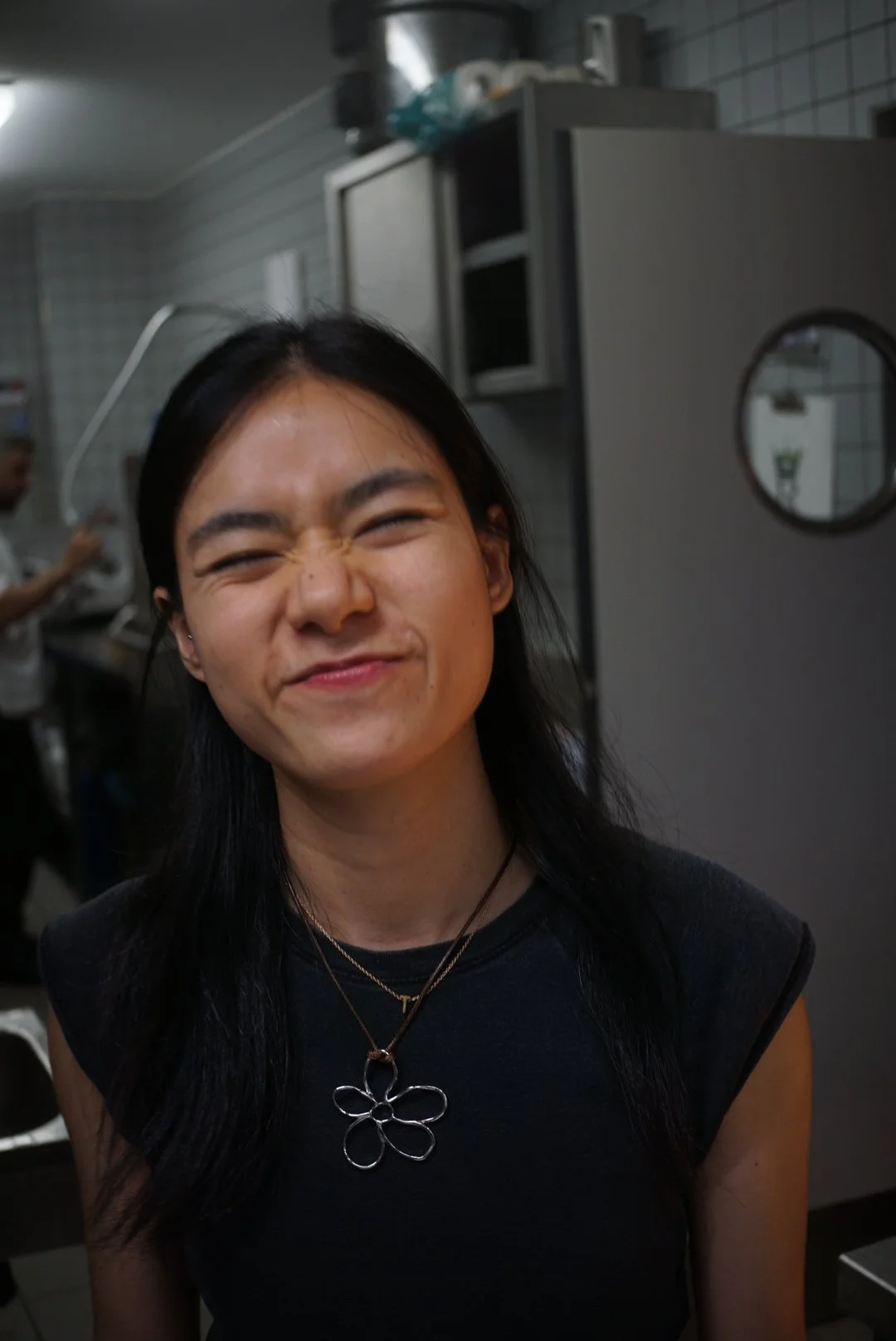 A woman with black hair making a scrunched face, standing in a commercial kitchen.
