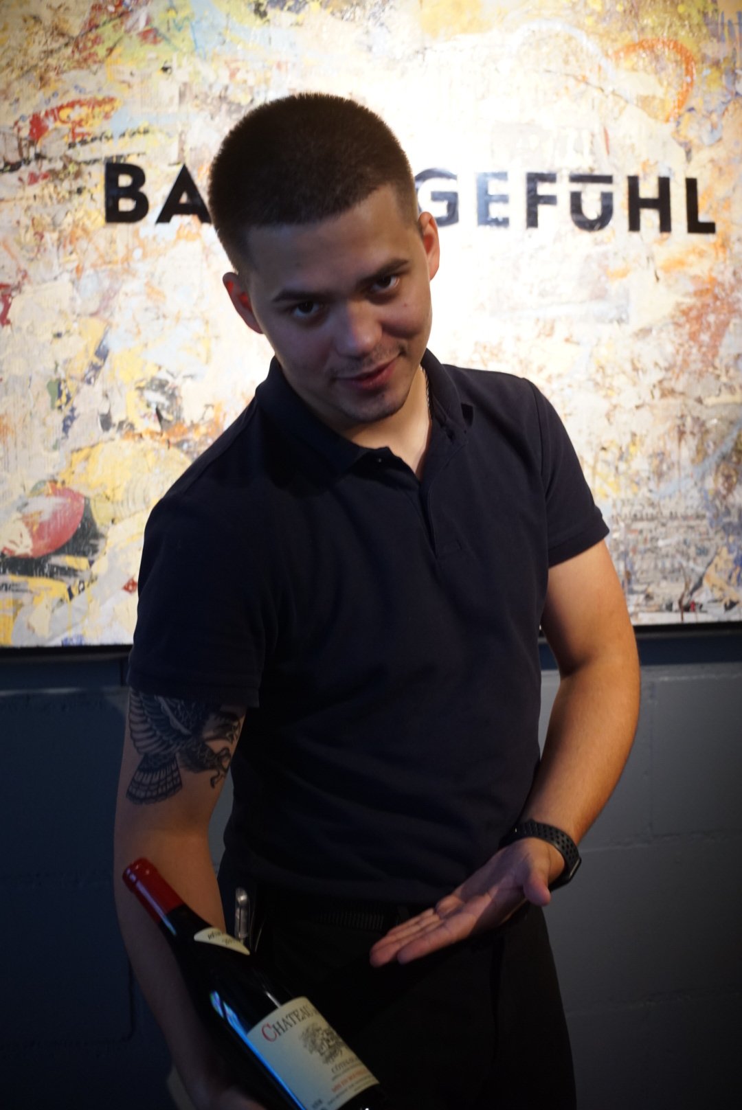 A young man with short dark hair and a tattoo of a bird on his right arm, holding a bottle of red wine, standing in front of a colorful abstract wall with partially visible text.