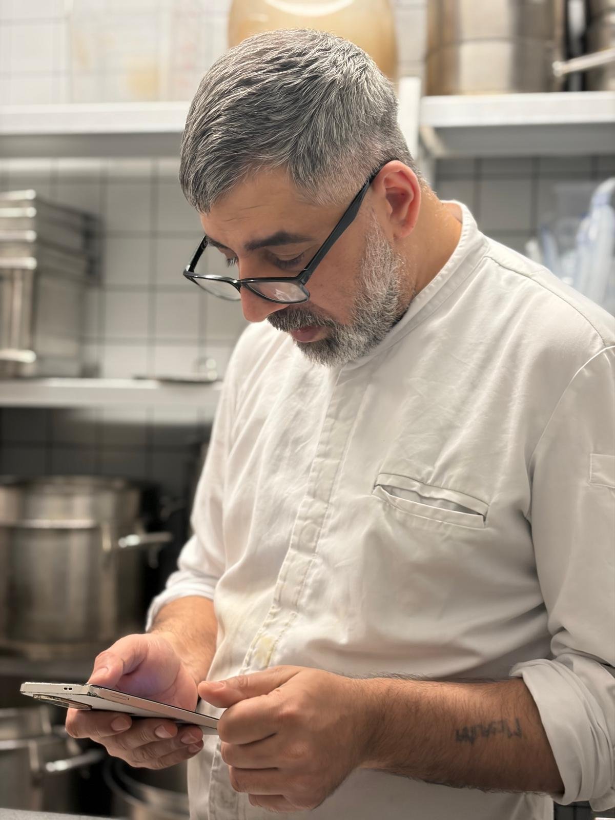 A male chef with gray hair and beard, wearing glasses, and a white chef jacket, standing in a commercial kitchen looking at his smartphone.
