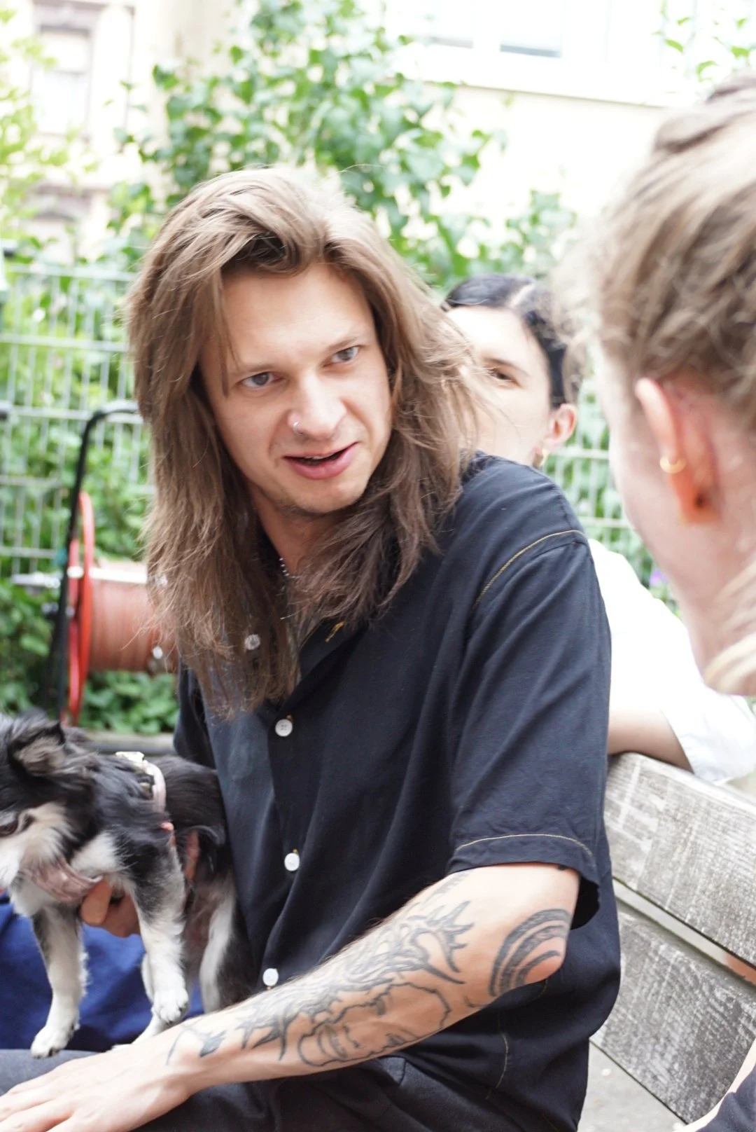 Young man with long hair, tattoos, and a nose piercing, holding a puppy while talking to a woman outside on a patio or garden.