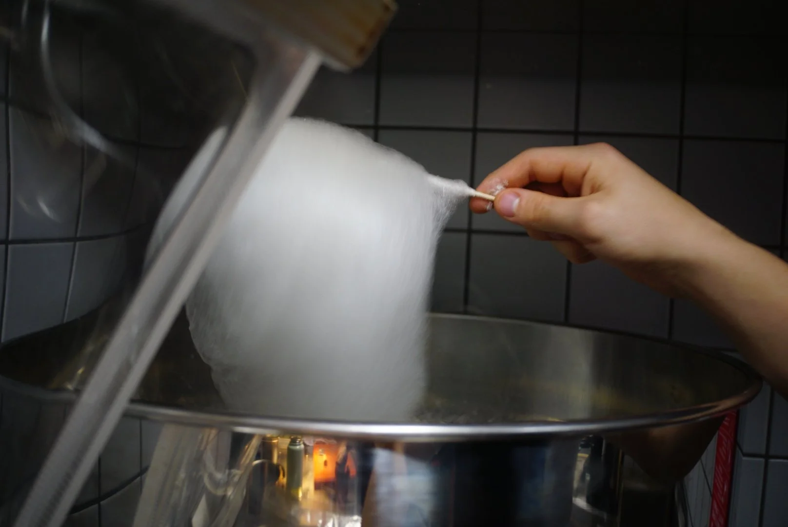 A person holding a cotton candy cone over a cotton candy machine, spinning out fluffy white cotton candy into a metal bowl, in front of a tiled wall.