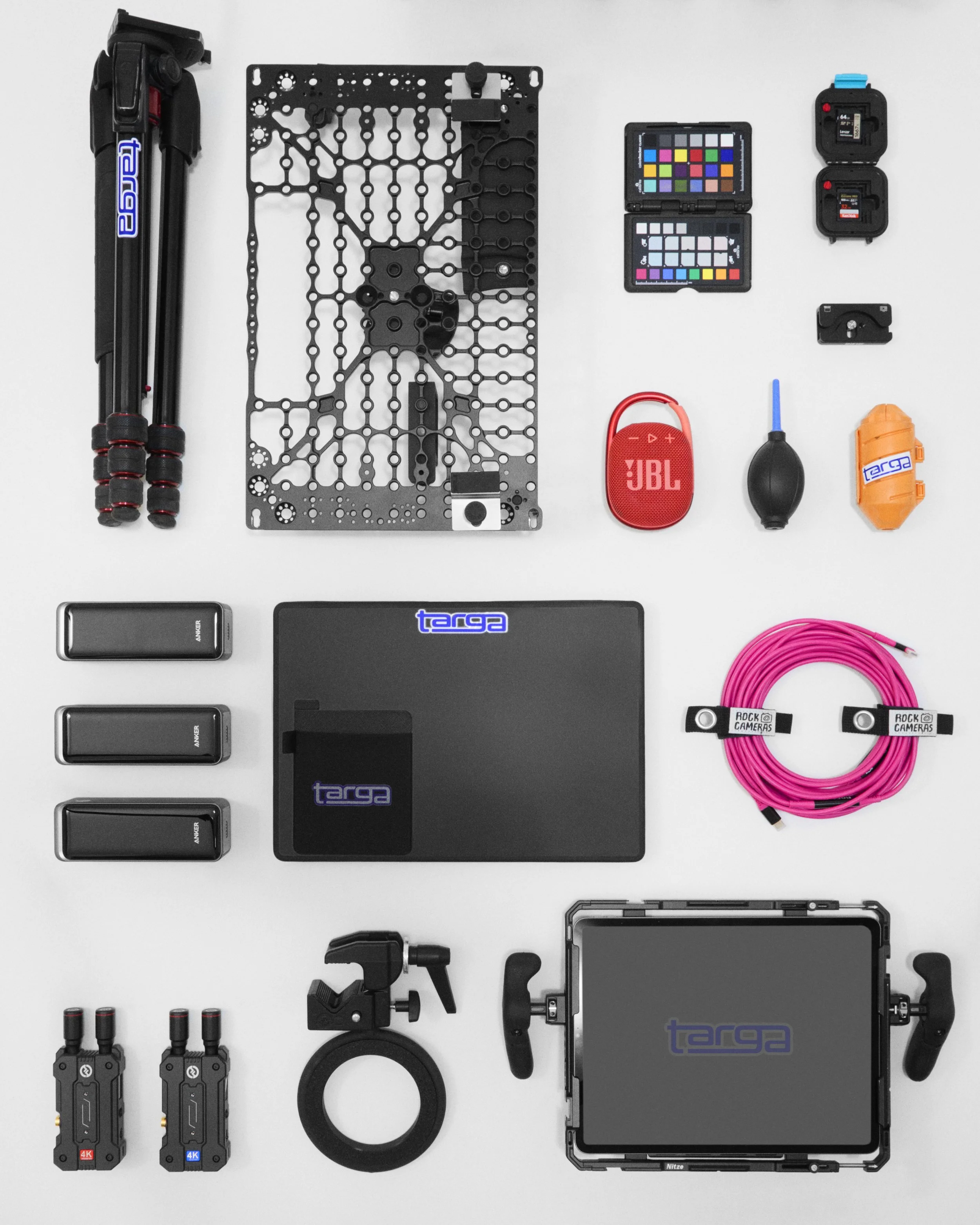 Varie attrezzature cinematografiche e di montaggio, tra cui luci, batterie, cavi, monitor, controller, e accessori, disposte ordinatamente su una superficie bianca.