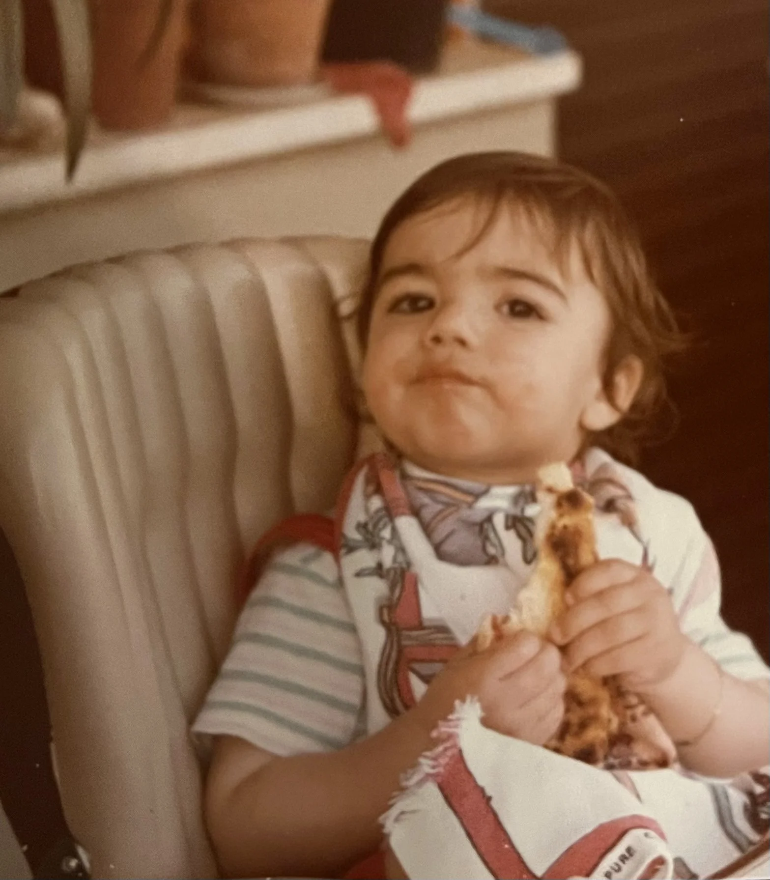 Bambino seduto su una sedia imbottita, che mangia una fetta di pizza, con cappello a maglia con disegni, in un ambiente domestico.