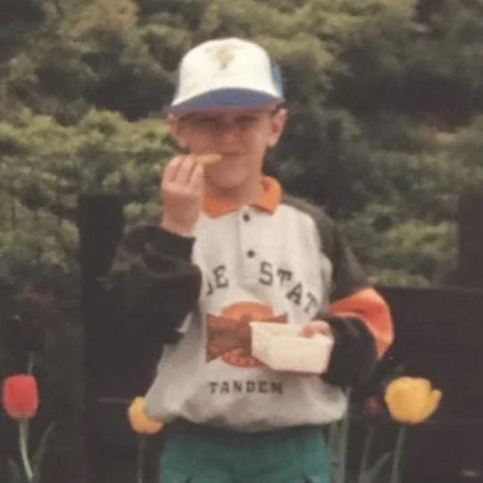 Bambino con cappello blu, maglietta con logo, che mangia uno snack in giardino.