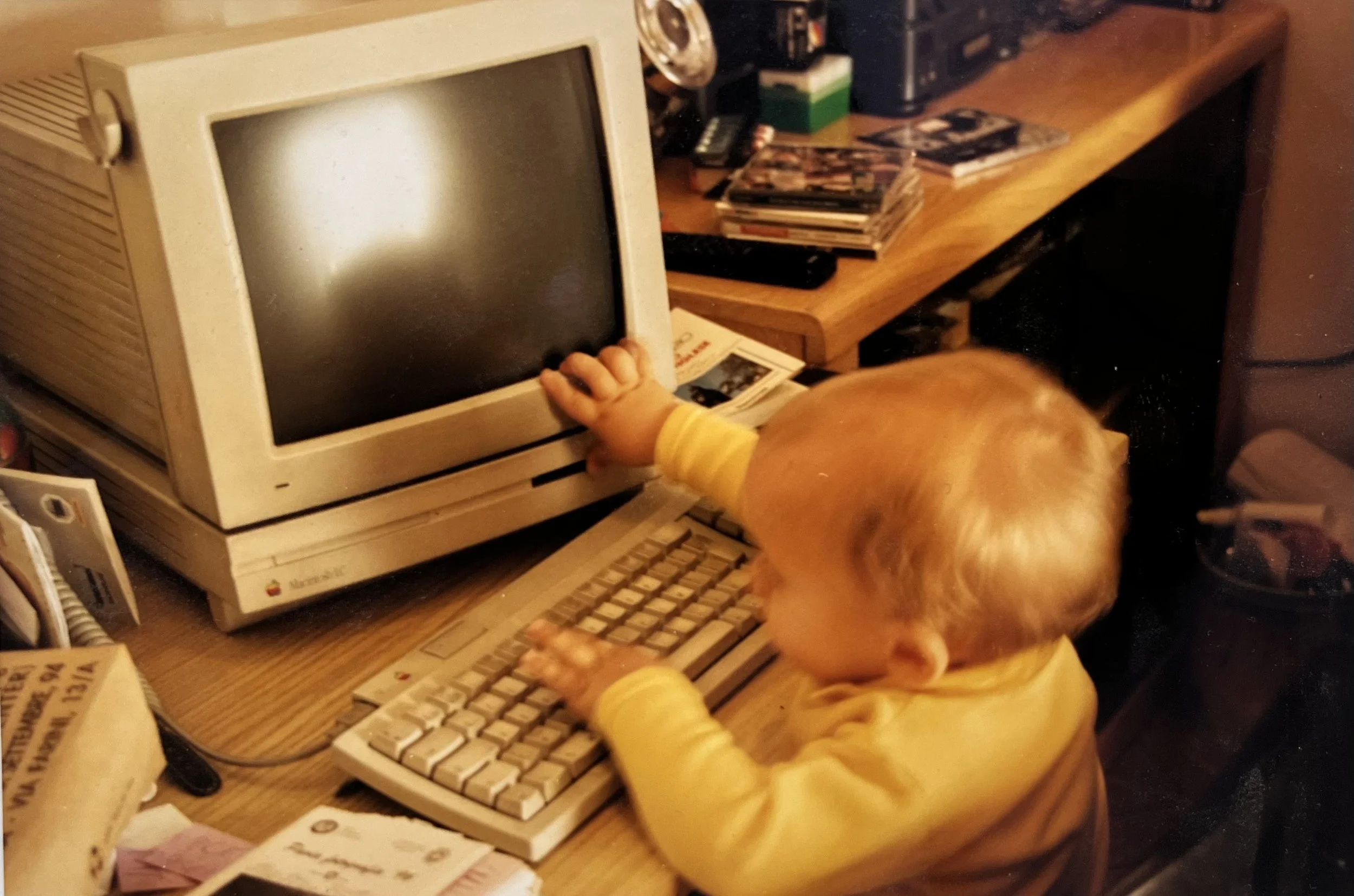 Bambino giovane che tocca uno schermo di un vecchio computer monitor di colore beige, posto sopra una tastiera che il bambino sta usando.