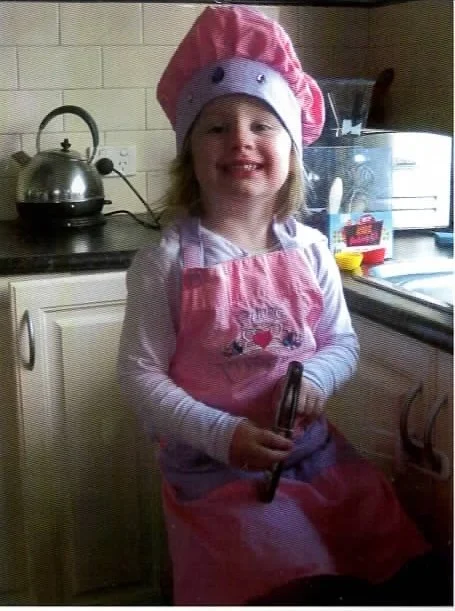 A young girl wearing a pink chef's hat and apron, holding a cake decorating tool, smiling in a kitchen.