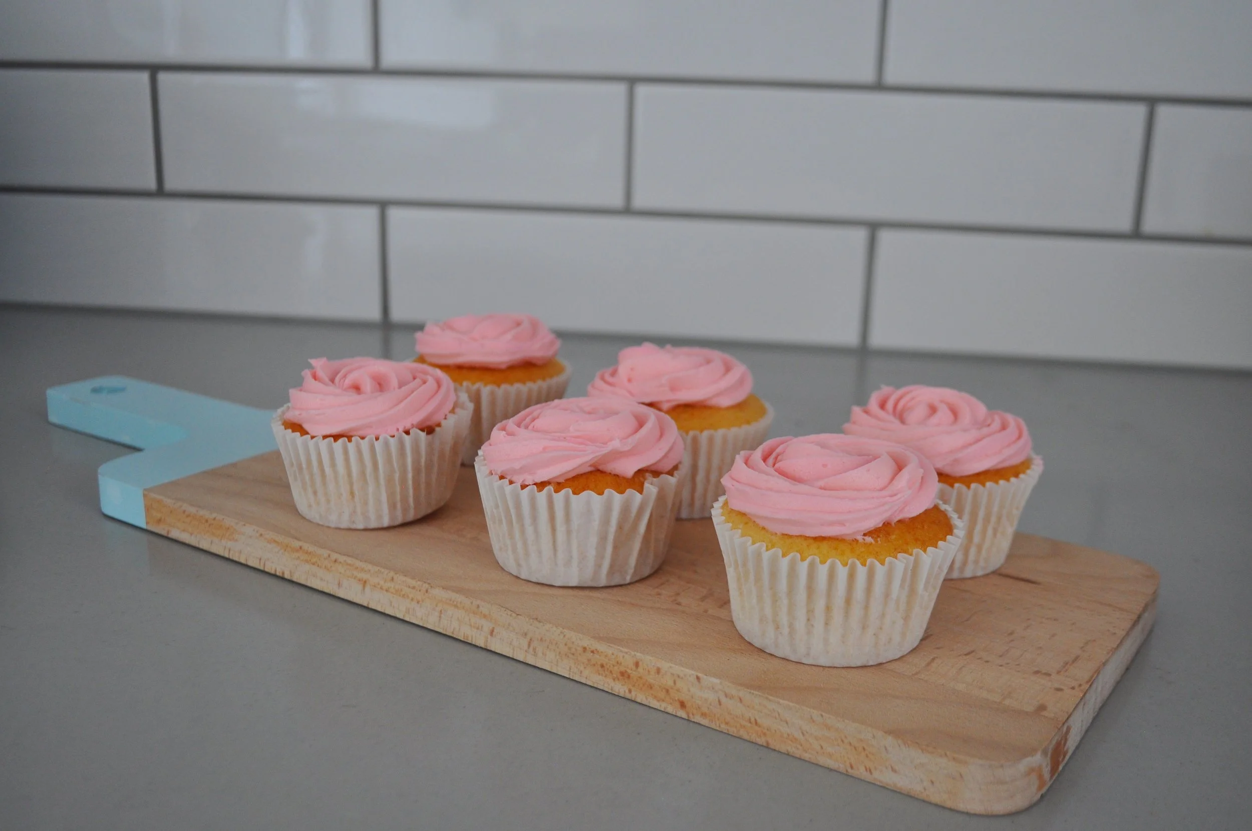 Six cupcakes with pink frosting swirls placed on a wooden cutting board on a gray countertop in front of a white tiled wall.