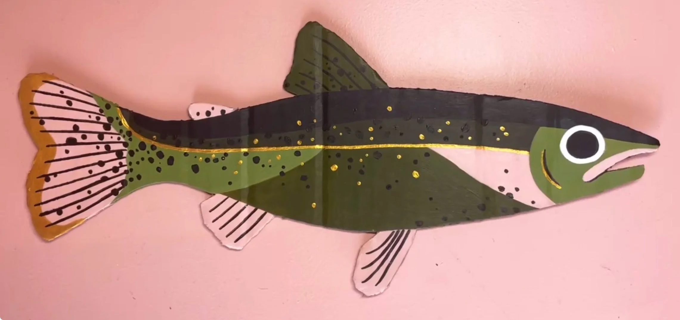 Colorful fish-shaped decoration with green, black, pink, and gold accents, displayed against a pink background.