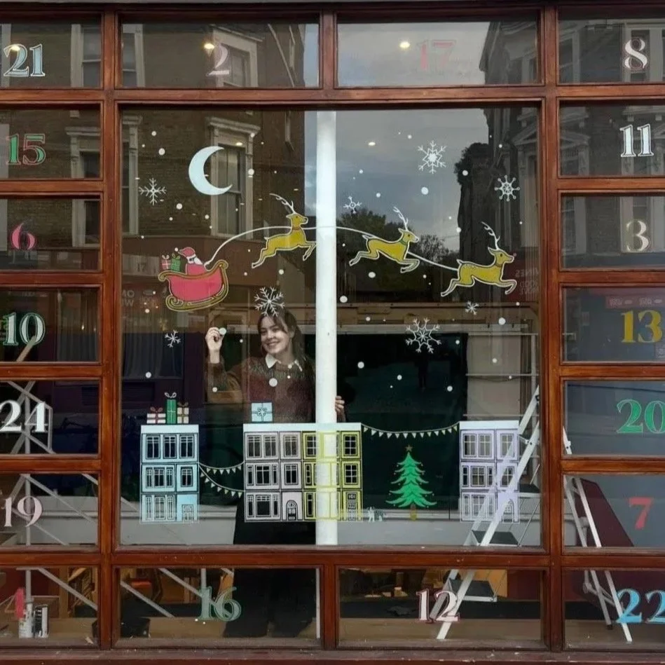 A Christmas-themed window decoration featuring a Santa with reindeer, snowflakes, a crescent moon, Christmas trees, and buildings, with an advent calendar numbered from 1 to 24.