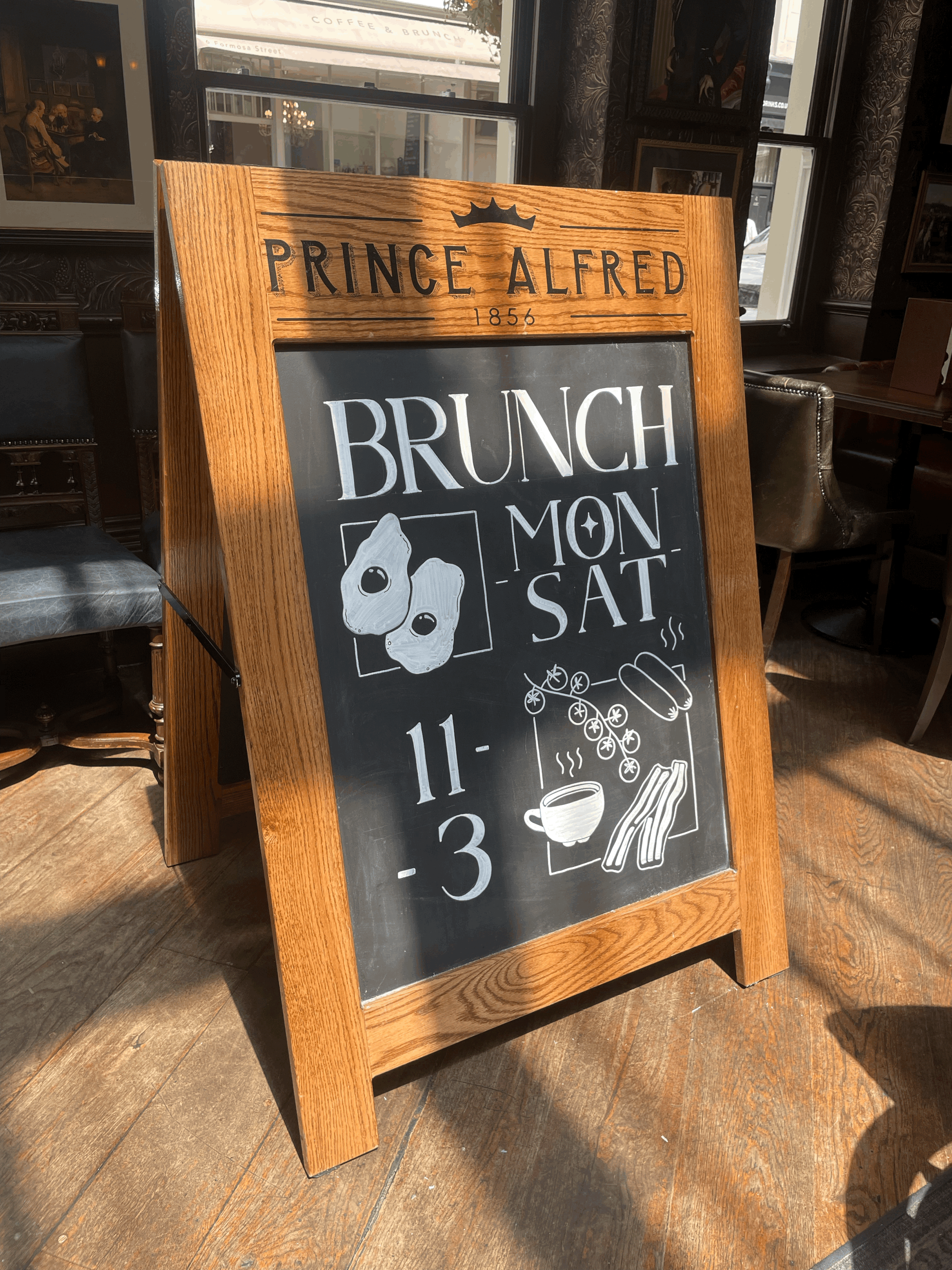 A wooden-framed chalkboard sign inside a pub with 'Prince Alfred 1856' at the top and 'Brunch Mon Sat 11-3' below, featuring illustrations of eggs, a hot beverage, and bacon.