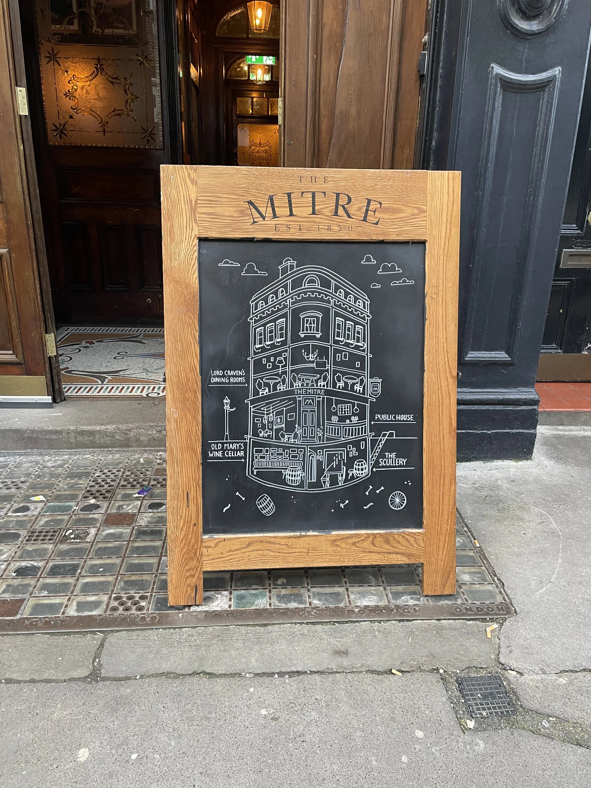 A wooden-framed chalkboard sign outside a pub, displaying a cartoon drawing of the building's interior layout with labels for different rooms, including a dining room, wine cellar, pub, and scullery.
