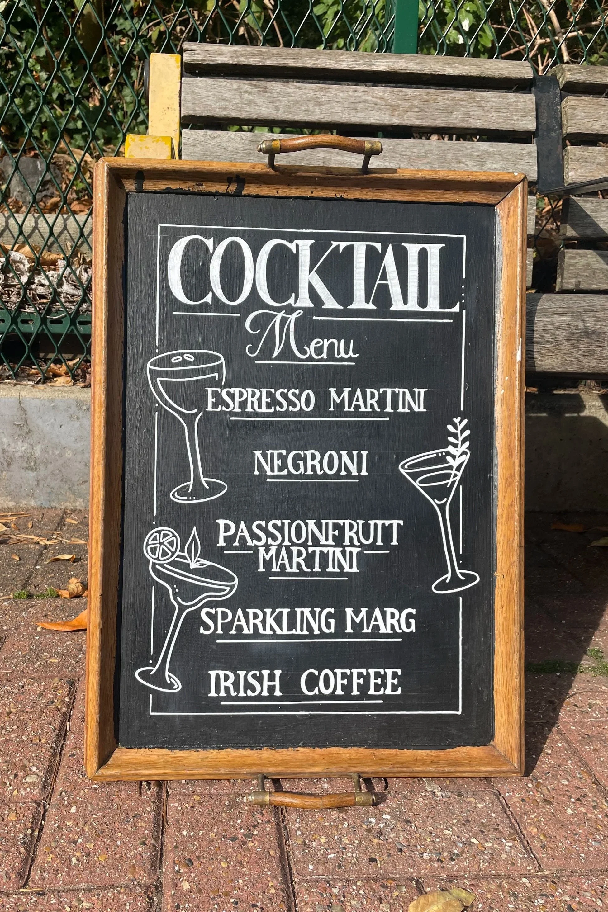 Chalkboard menu outside a bar or café listing cocktails: Espresso Martini, Negroni, Passionfruit Martini, Sparkling Margarita, Irish Coffee, with illustrations of cocktails on a black background framed with wood.