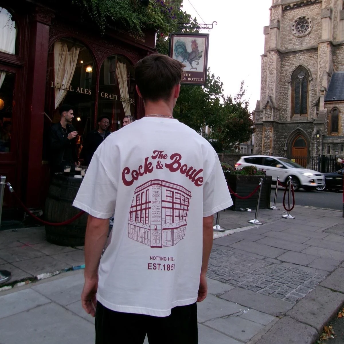 A person wearing a white T-shirt with red text and illustration on the back, standing on a city sidewalk near a bar and a historic church, with cars passing by.