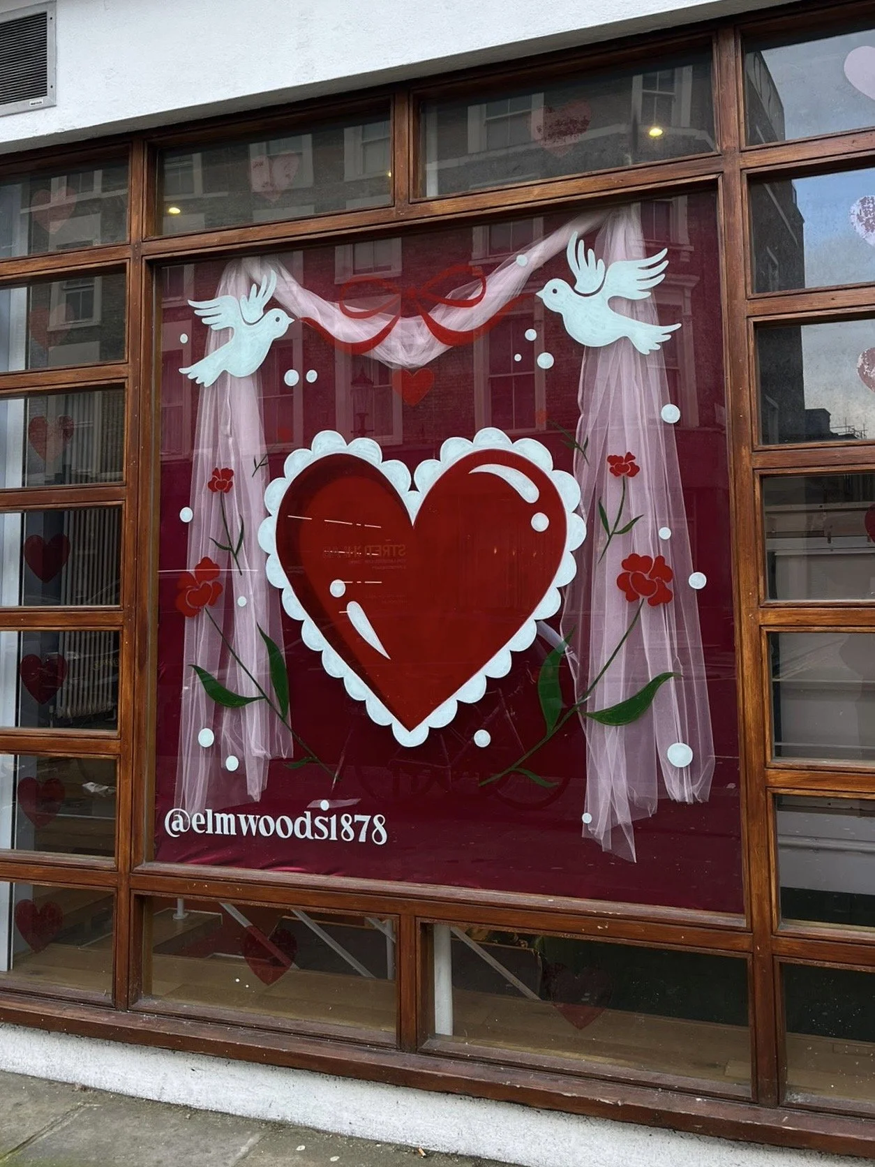 Decorative window display with a large red heart, white doves, pink curtains, red flowers, and an Instagram handle '@elmwoods1878'.