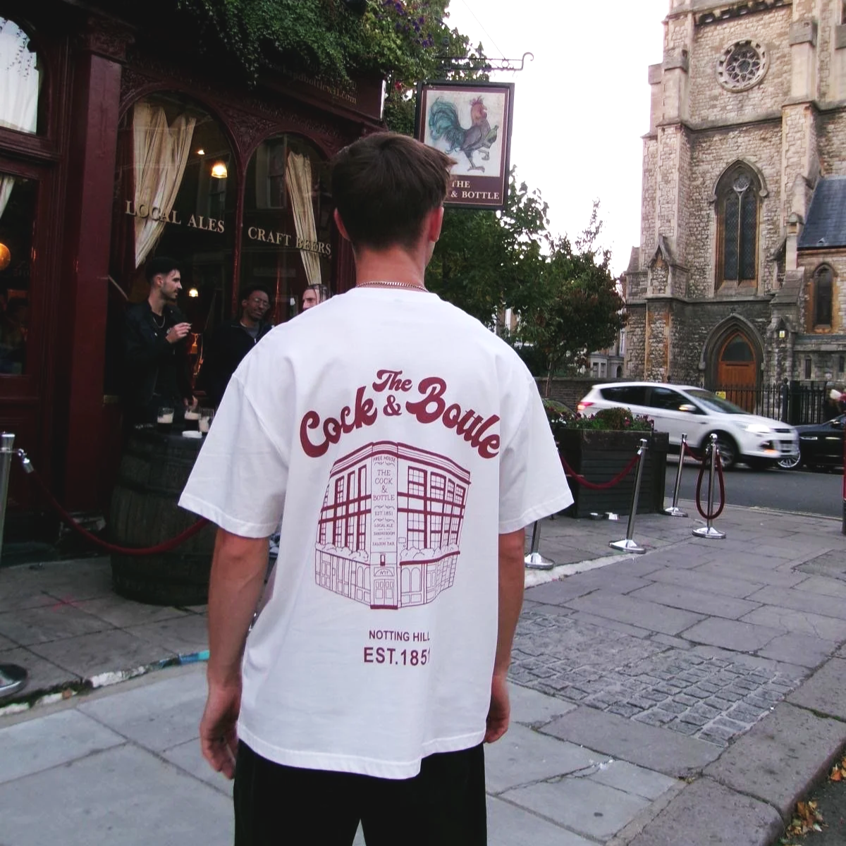 A young man standing on a city sidewalk facing away, wearing a white T-shirt with red text and illustration for The Cock & Bottle pub, with an old church and a few people talking outside a bar or pub in the background, and a car passing on the street.