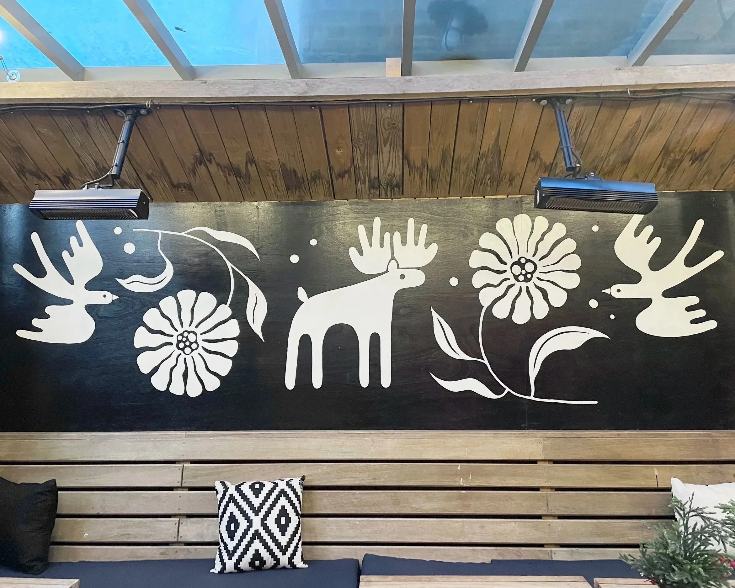 A wooden bench with two black cushions and black-and-white patterned cushions, in front of a black wall with white floral and animal illustrations and a glass ceiling above.
