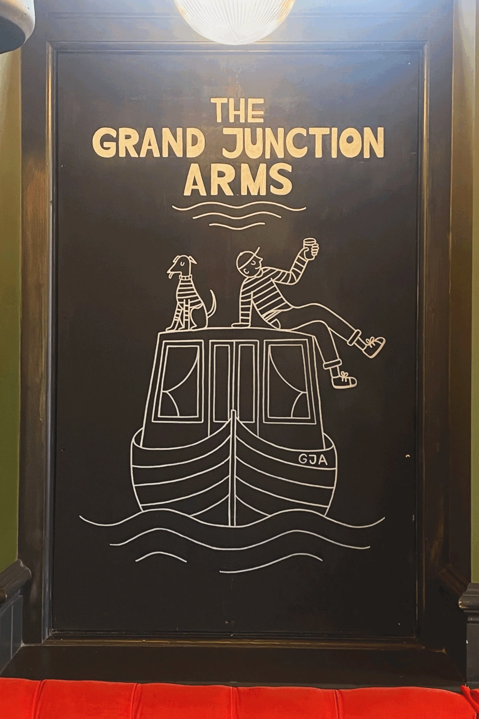 Chalkboard sign with white illustrations of a boat, a person sitting on top of the boat with a dog, and the text 'The Grand Junction Arms' at the top.