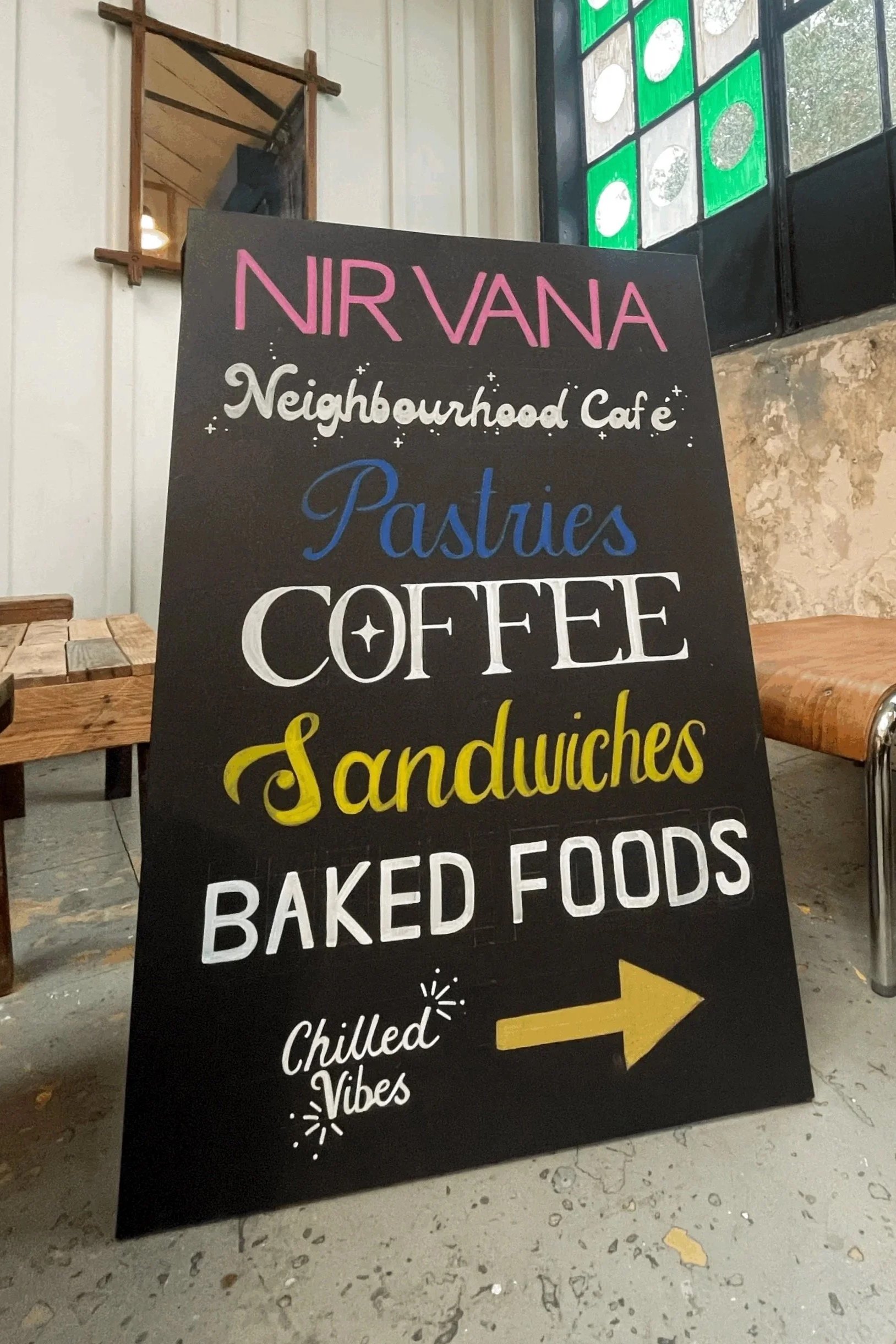 A black chalkboard-style sign with colorful handwritten text displaying menu items such as Nirvana neighborhood cafe, pasties, coffee, sandwiches, baked foods, and chilled vibes, with a yellow arrow pointing to the right.