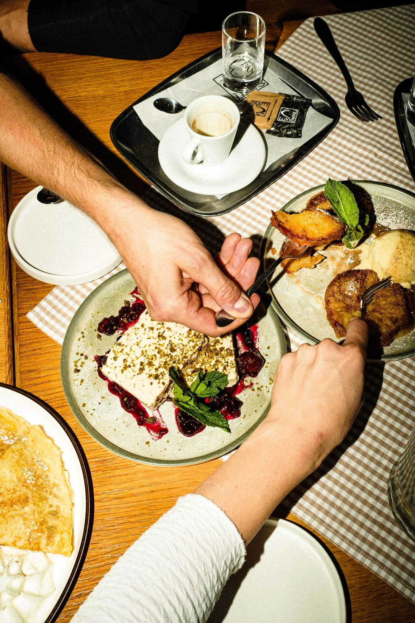Hands serving dessert on a table with plates of food, a coffee cup, spoon, fork, and empty glass.