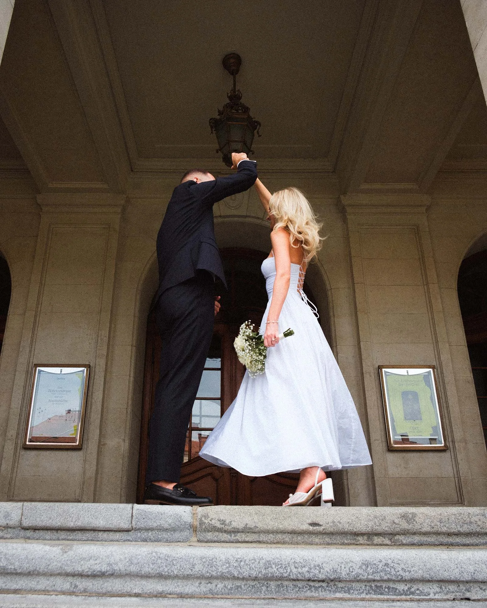 A couple dressed in wedding attire dancing on steps in front of a building with stone walls and large wooden doors. The man is in a black suit and the woman in a white wedding dress holding a small bouquet of flowers.
