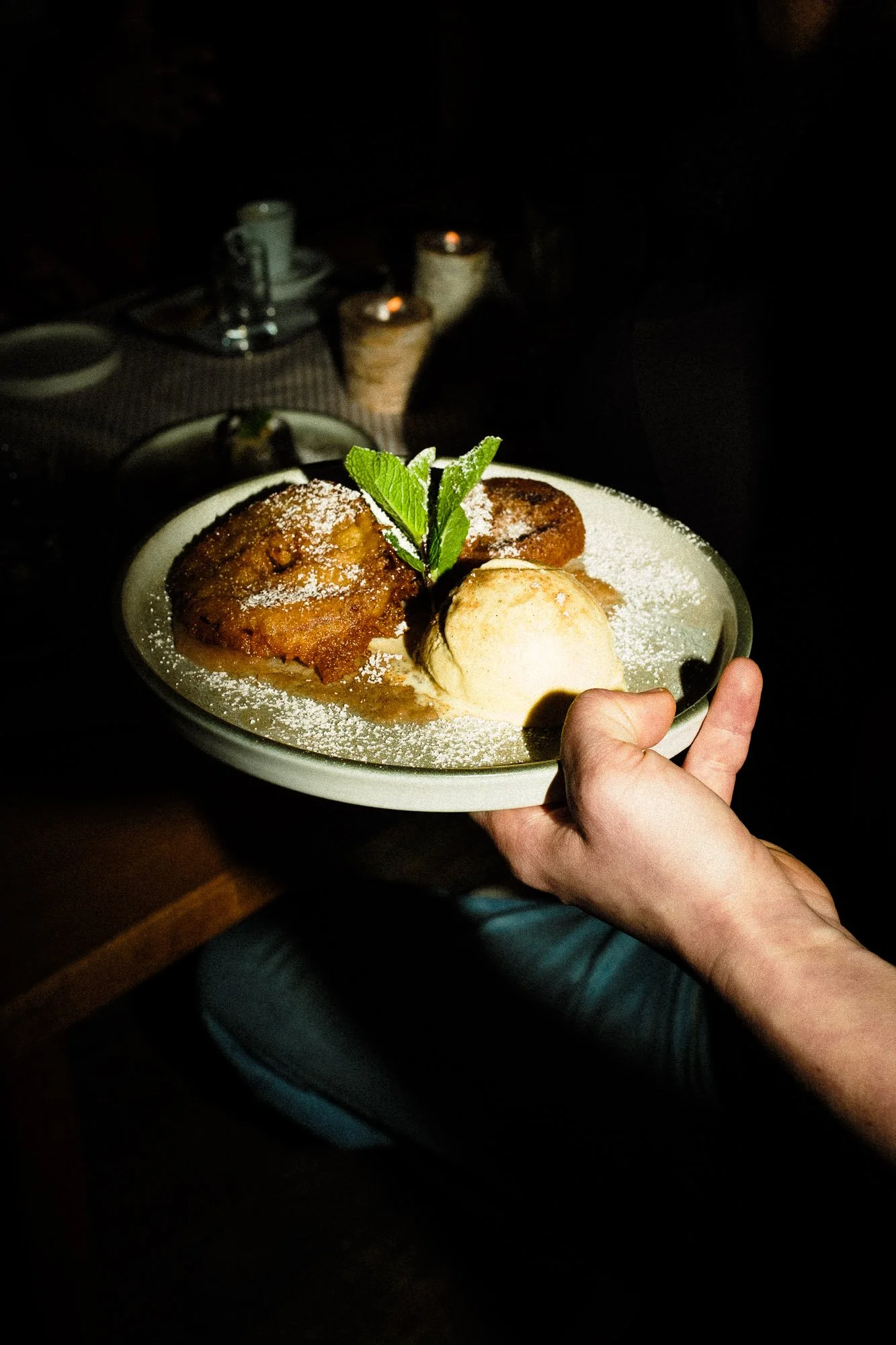 A hand holding a white plate with apple cobbler topped with powdered sugar, a scoop of vanilla ice cream, and a sprig of mint, in a dimly lit setting.
