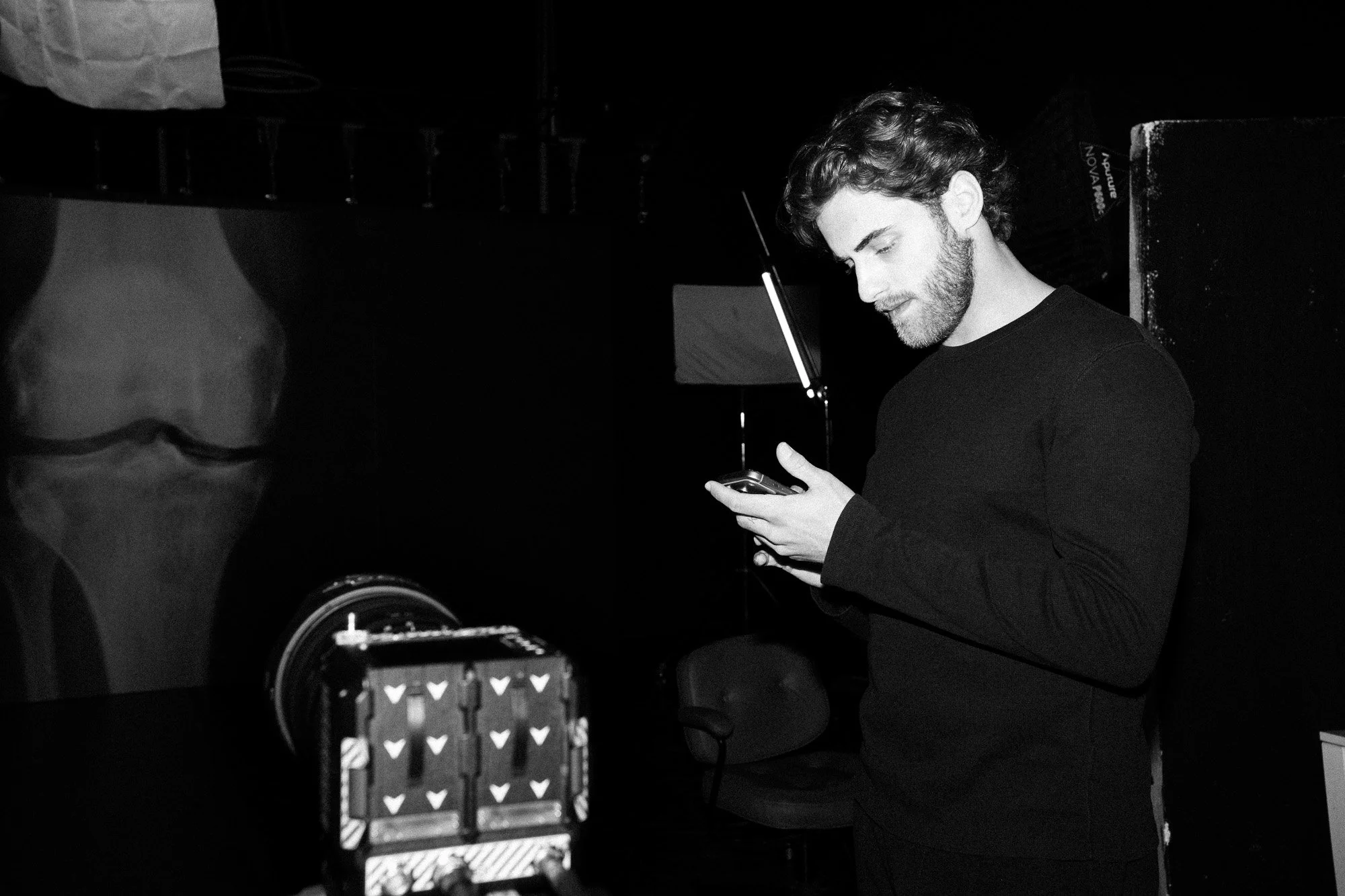 A man with curly hair and a beard standing in a dark room, looking at his phone.
