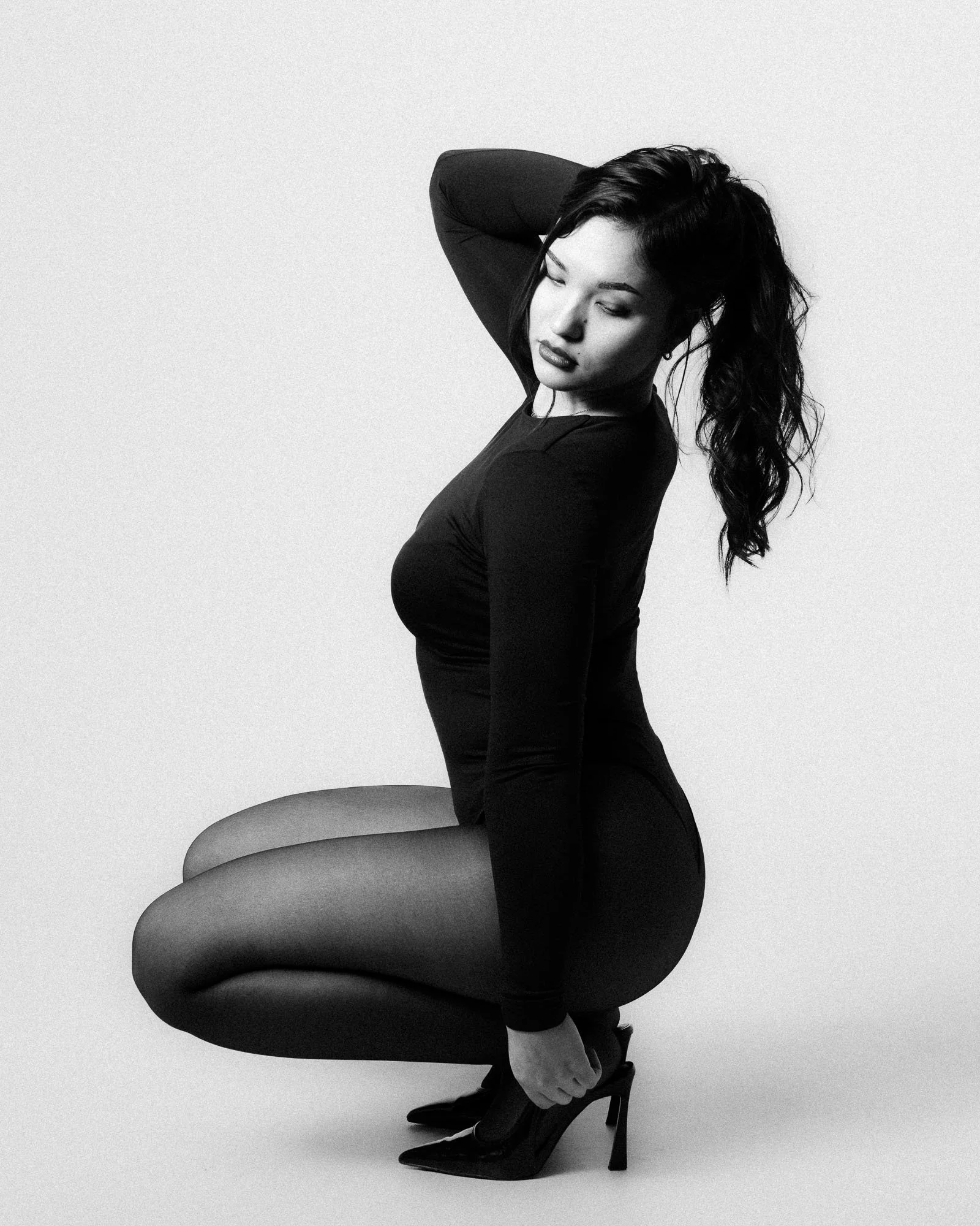 A black and white photo of a woman with long, wavy hair, squatting down while holding her high-heeled shoes and looking down with her eyes closed.
