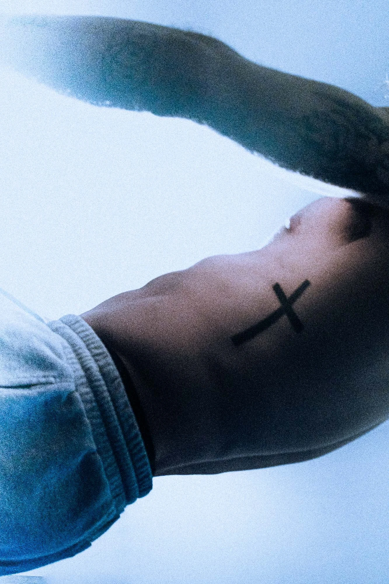 Close-up of a person's side, showing a black cross tattoo on their neck, with part of their face and a sleeve visible, and a light blue background.