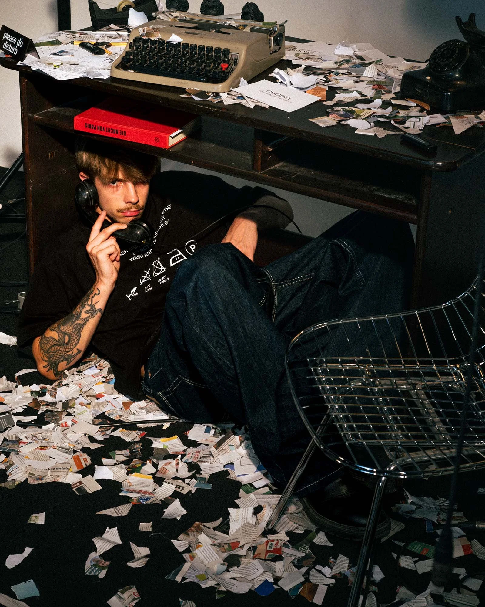 A young man with tattooed arm lying on the floor under a cluttered desk, holding a phone to his ear, surrounded by torn paper and documents, with an old typewriter, telephone, and scattered papers on the desk.