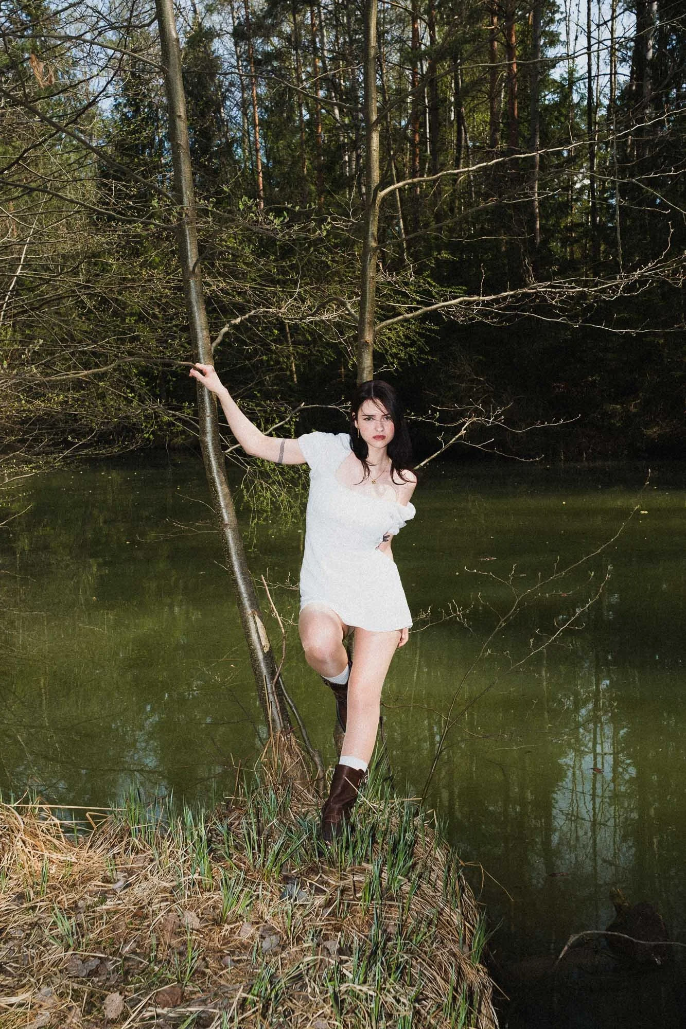 A woman with dark hair in a white dress and brown boots standing on a small grassy island by a pond, holding onto a thin tree trunk with one hand and looking serious.