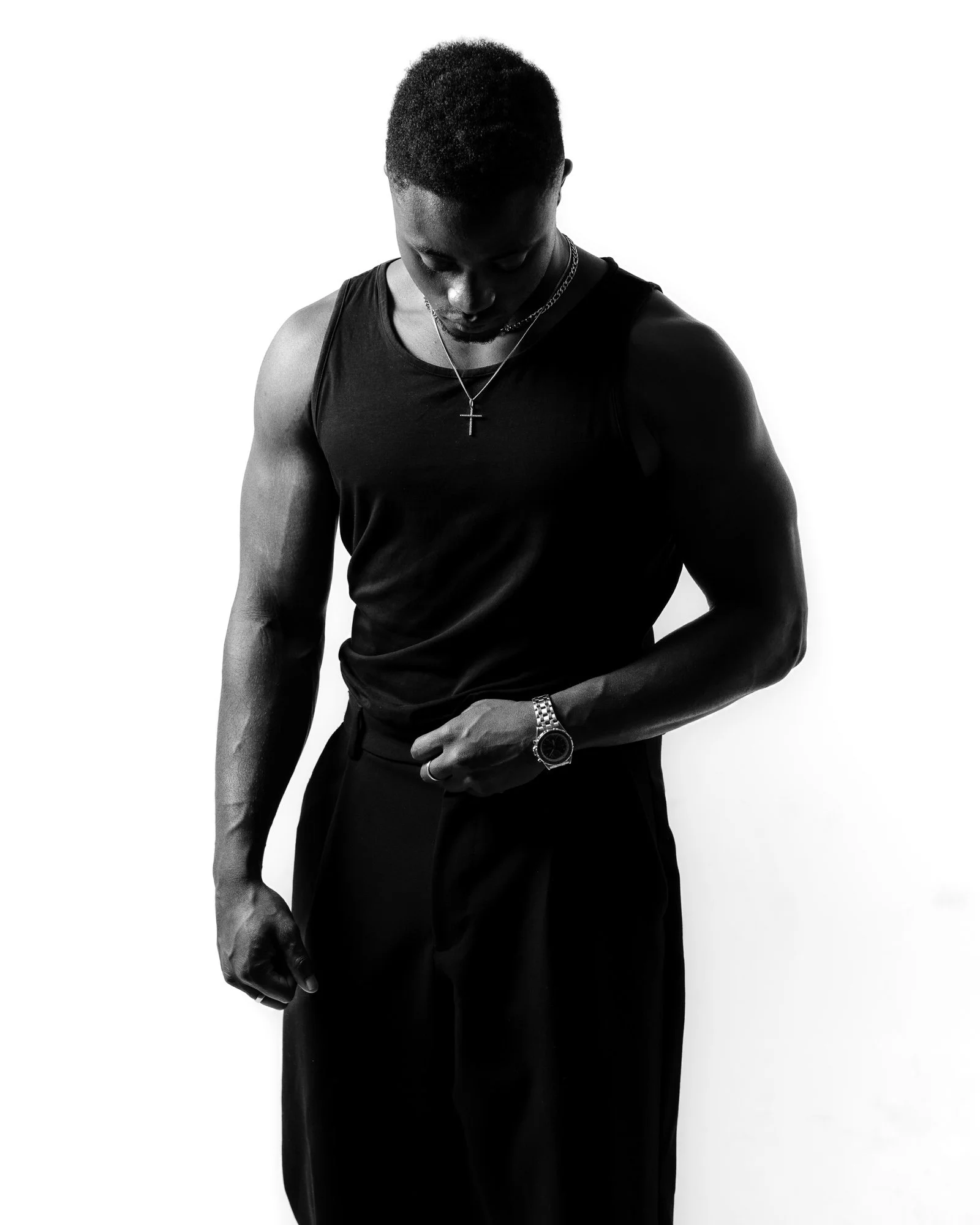 Black and white photo of a man looking down, wearing a sleeveless shirt, watch, and a cross necklace, standing against a plain white background.