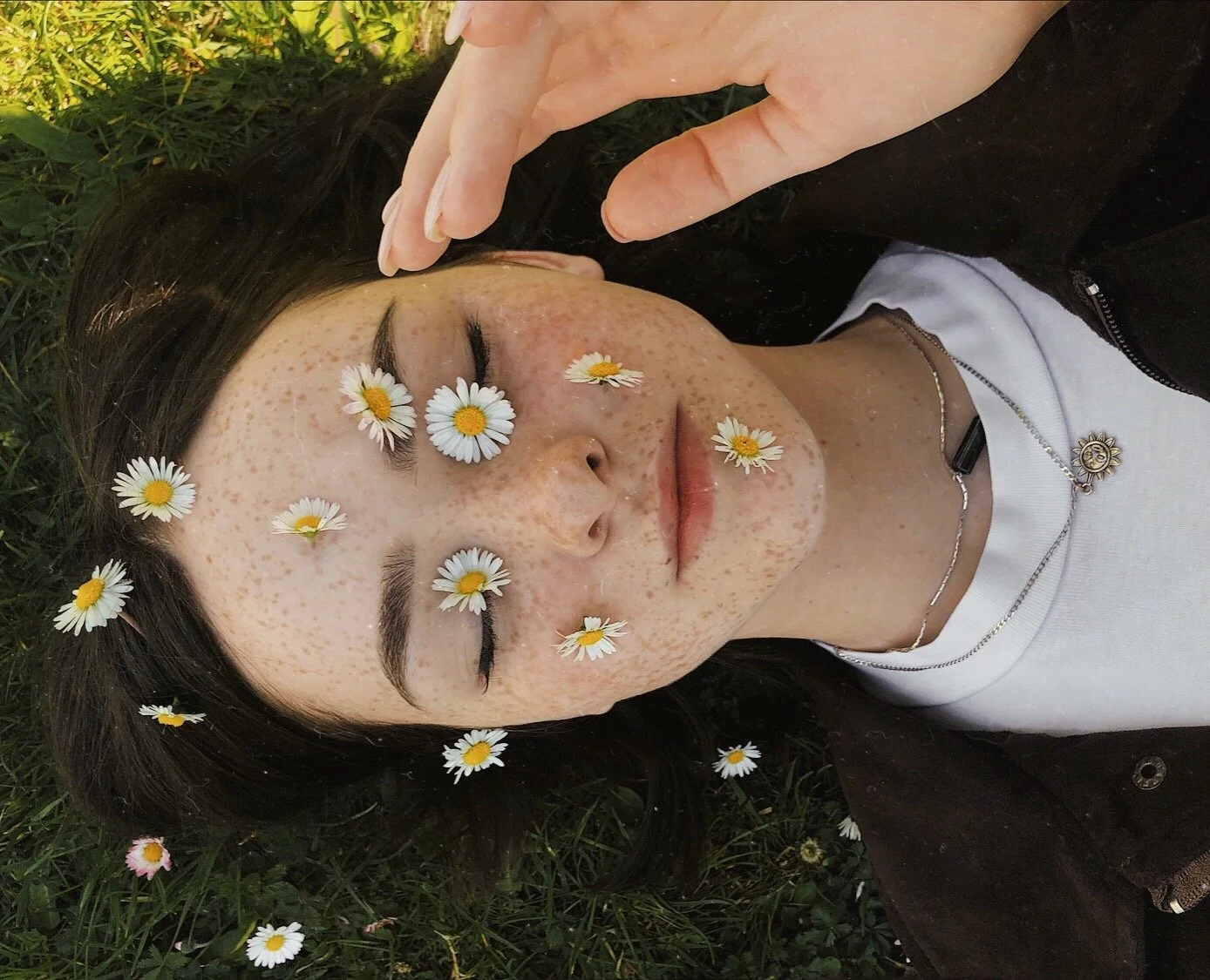 A woman with freckles and dark hair lying on grass with flowers on her face, eyes closed, and a hand near her forehead.
