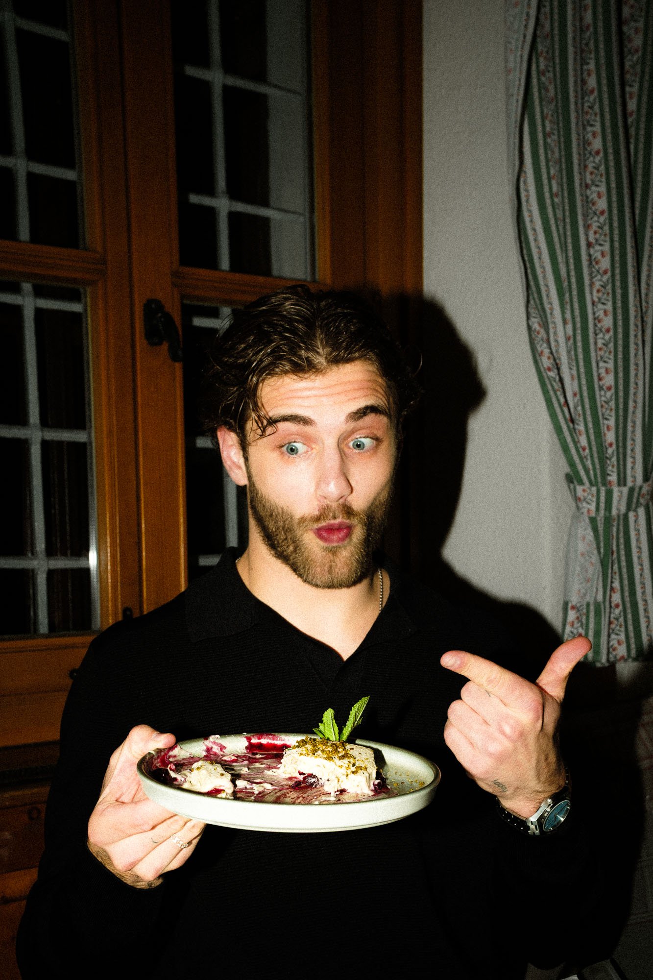 A man with brown hair and beard, wearing a black shirt, is holding a plate with a dessert garnished with a green herb, and pointing at it with a surprised expression. The background shows a wooden-framed window, part of a curtain with green and red s
