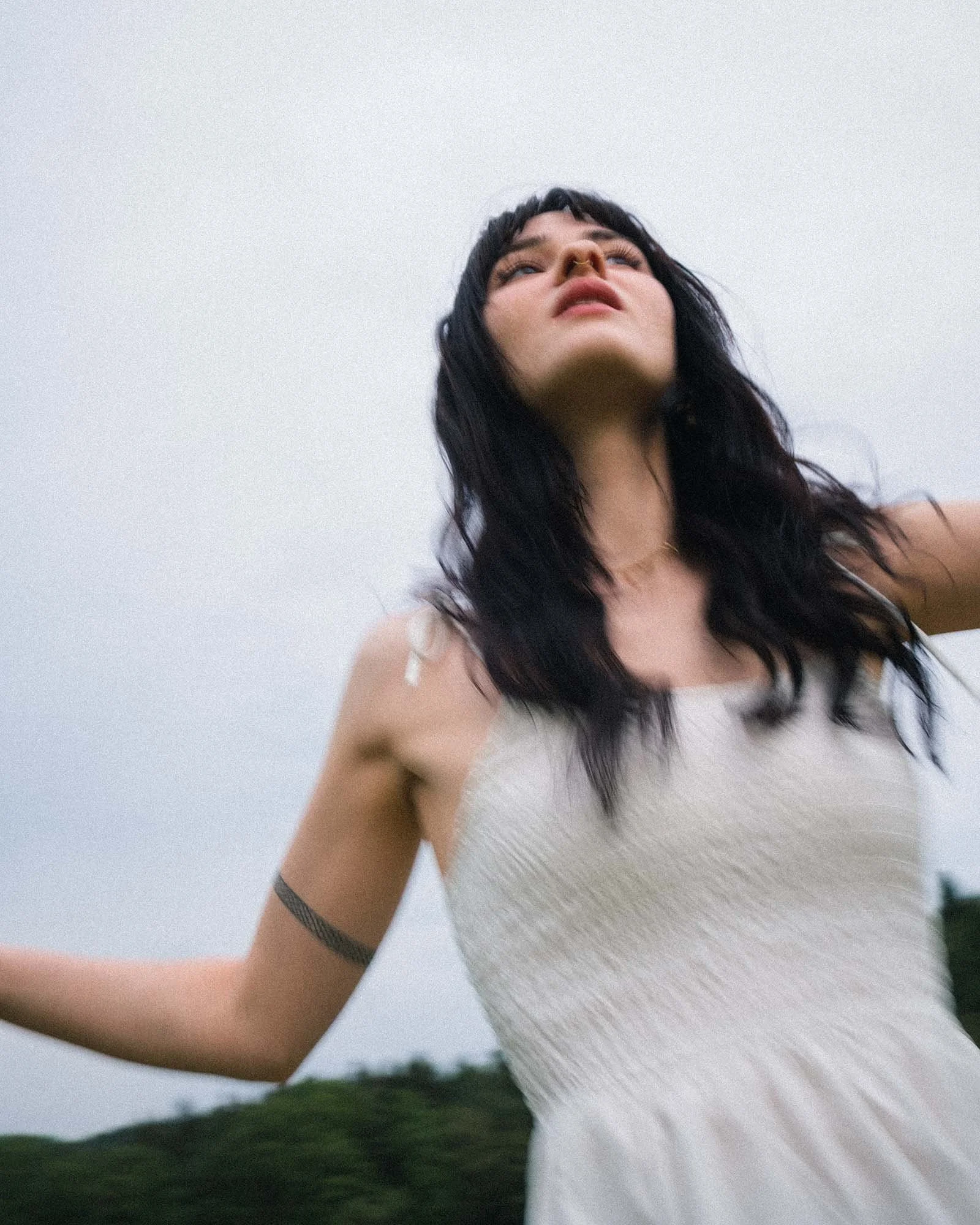 A woman with dark wavy hair in a white sleeveless dress standing outdoors against an overcast sky and greenery.