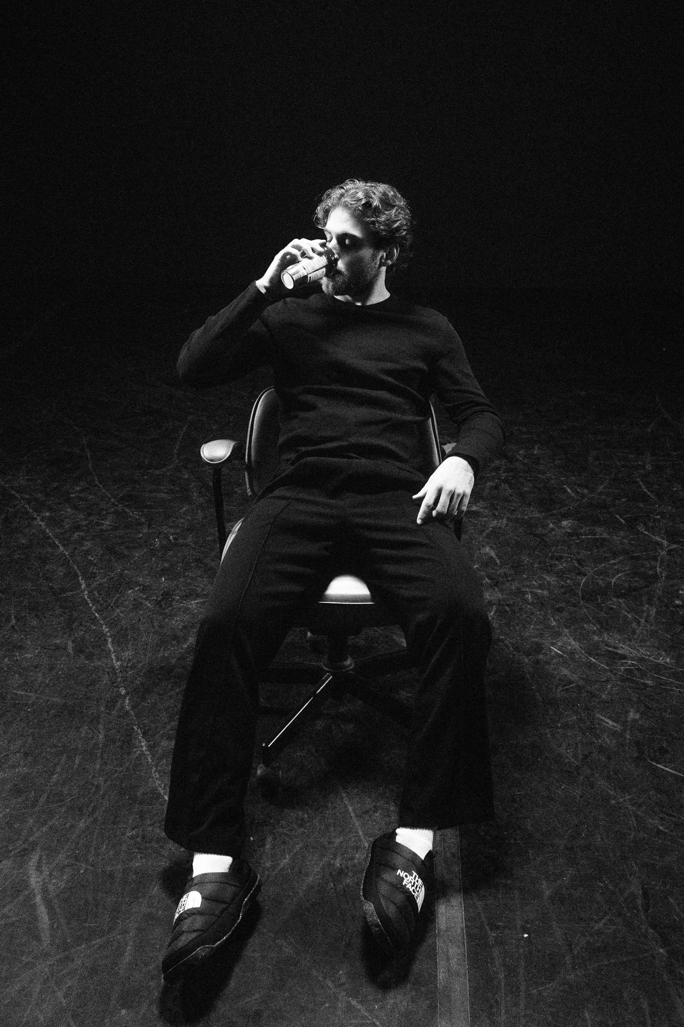 A man sitting on a chair on a dark stage, drinking from a can, with casual attire and sneakers, in a black and white photograph.