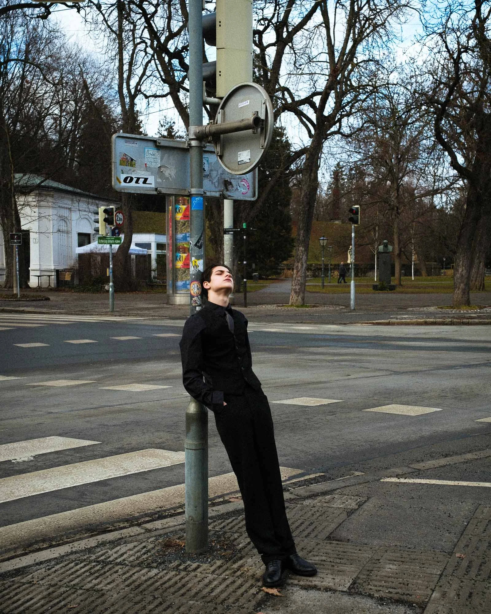 A person in black clothing leaning against a street lamp on a city street, looking up with eyes closed. There are traffic lights and trees in the background.