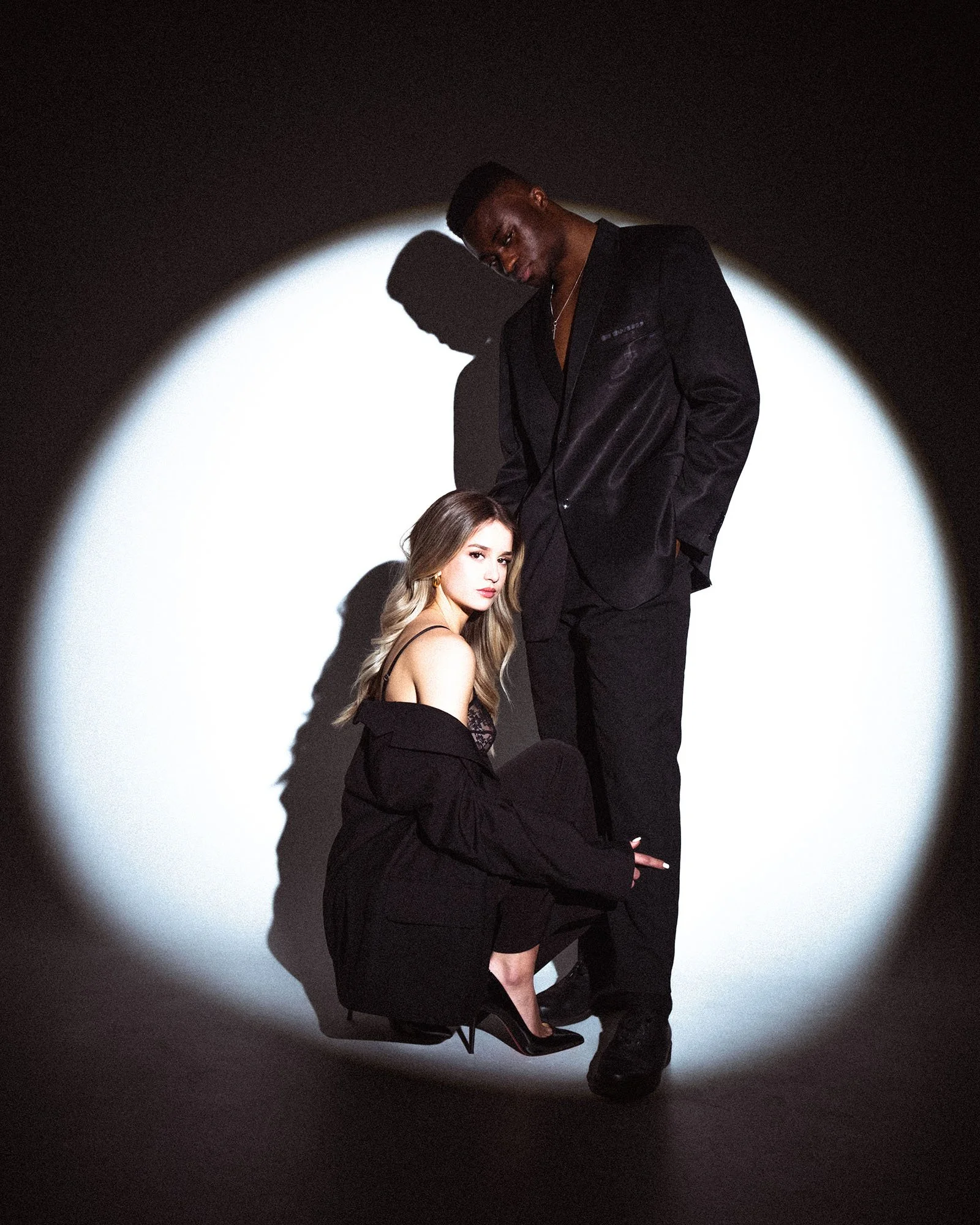 A man and woman posing together in a dark setting, illuminated by a circular spotlight, both dressed in black clothing.