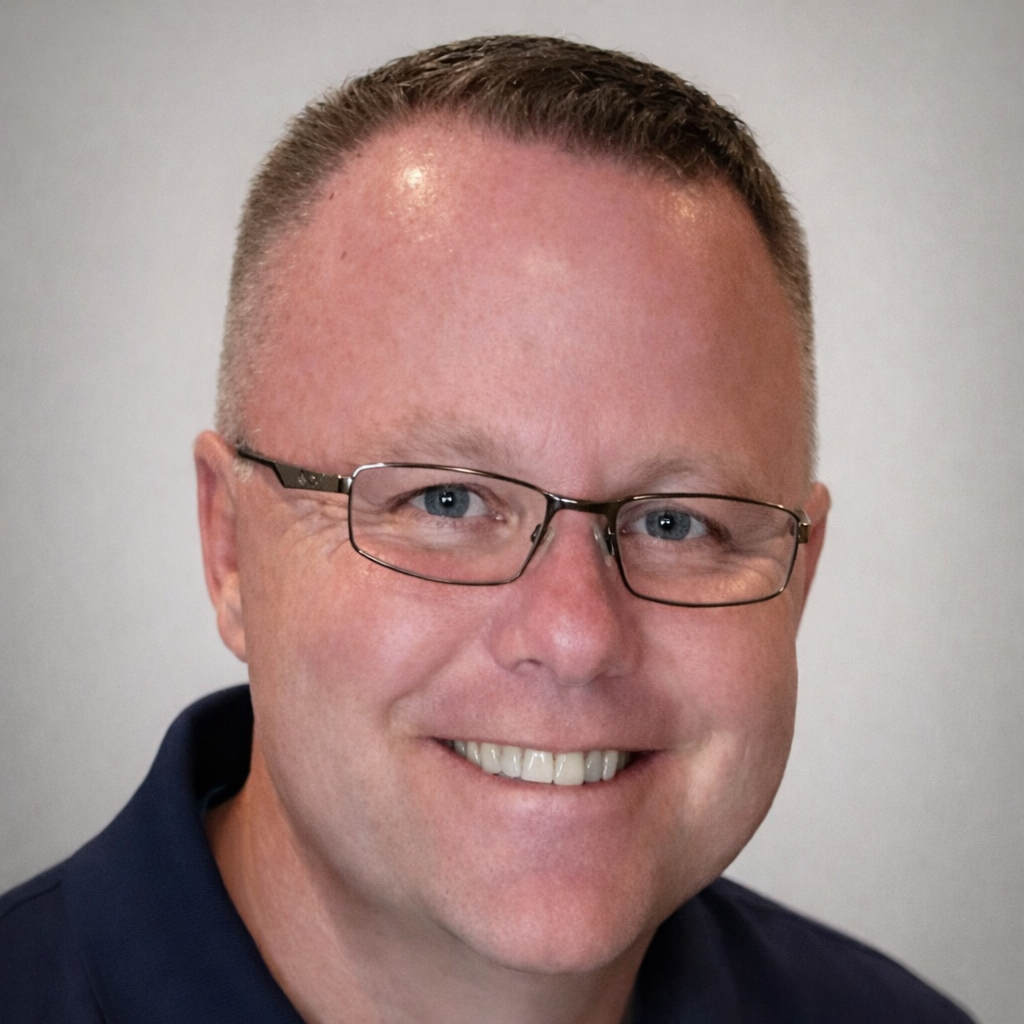 A smiling man with short brown hair, glasses, and blue eyes, wearing a dark blue shirt centrally shot against a plain light background.