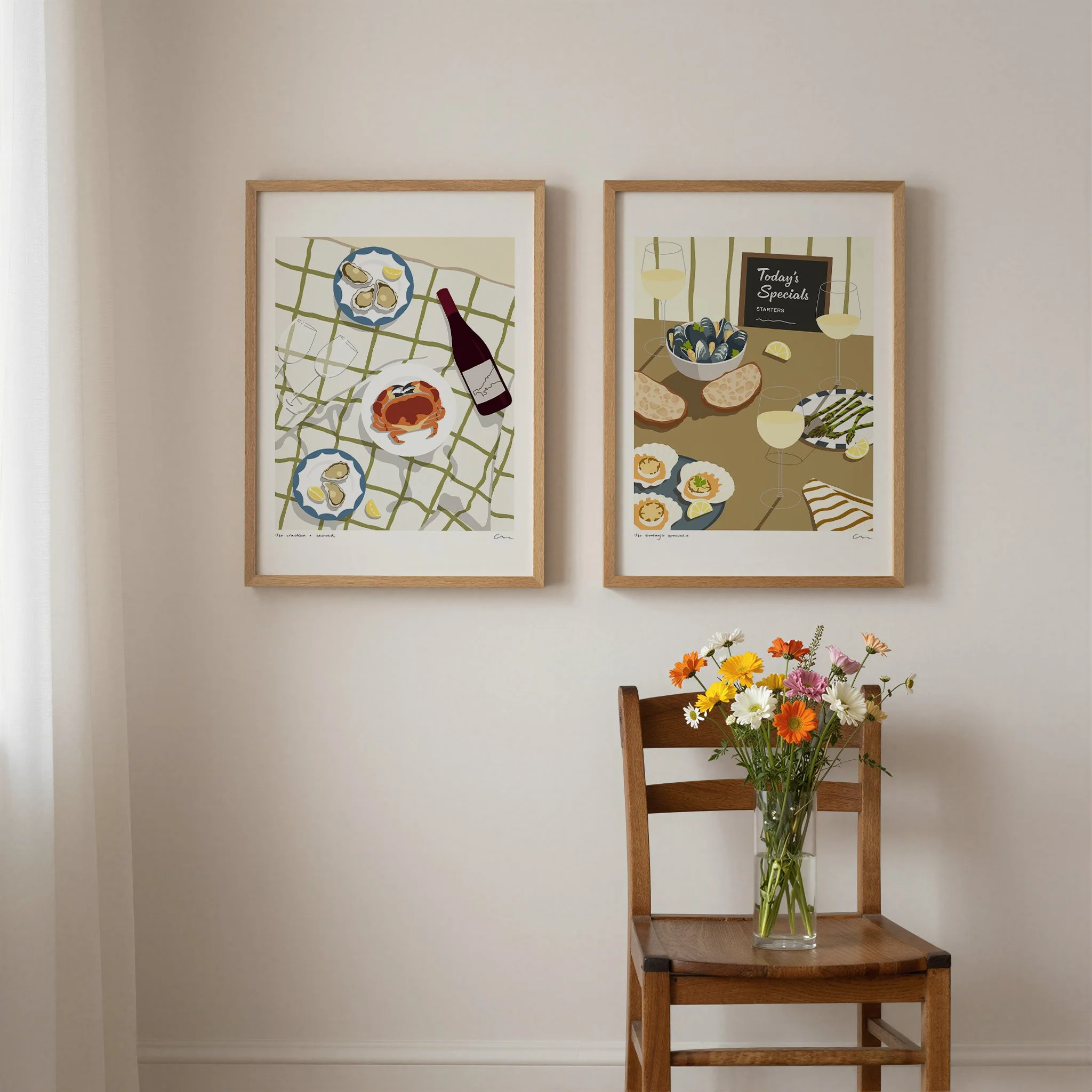 Two framed colorful illustrations of seafood dishes hanging on a white wall. A wooden chair with a vase of mixed flowers is in front of the wall.