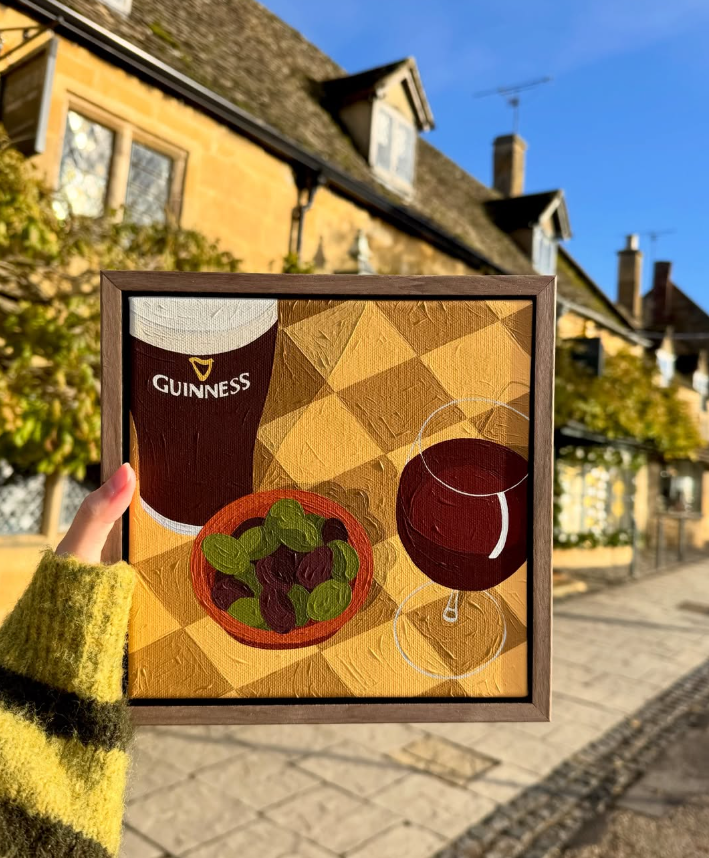 Painting of a Guinness beer, a glass of red wine, and a bowl of olives on a checkered tablecloth.