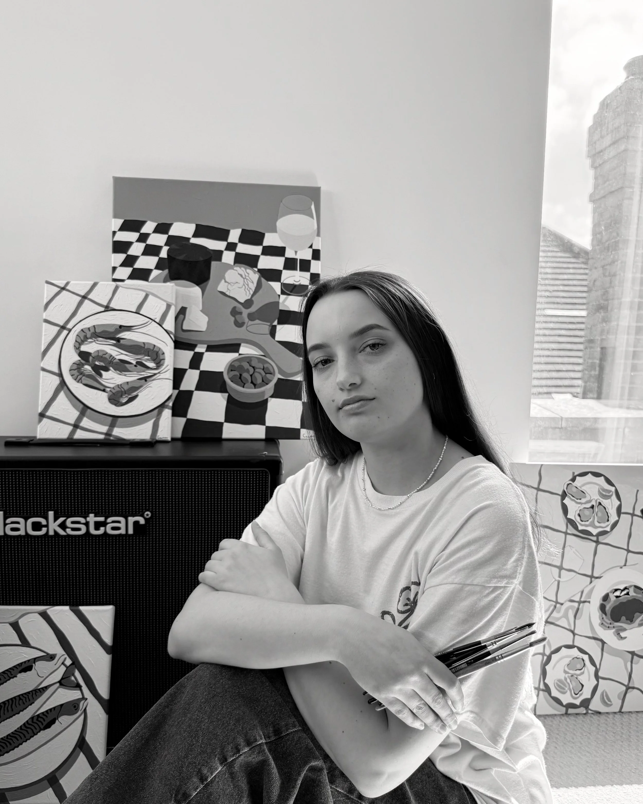 A woman with long dark hair and a light-coloured shirt sitting in front of a selection of paintings.
