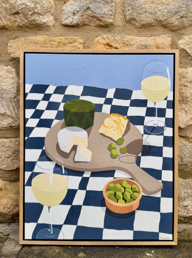 Painting of a table with a cheese board, a bowl of olives, and white wine on a checkered tablecloth against a Cotswold stone wall.