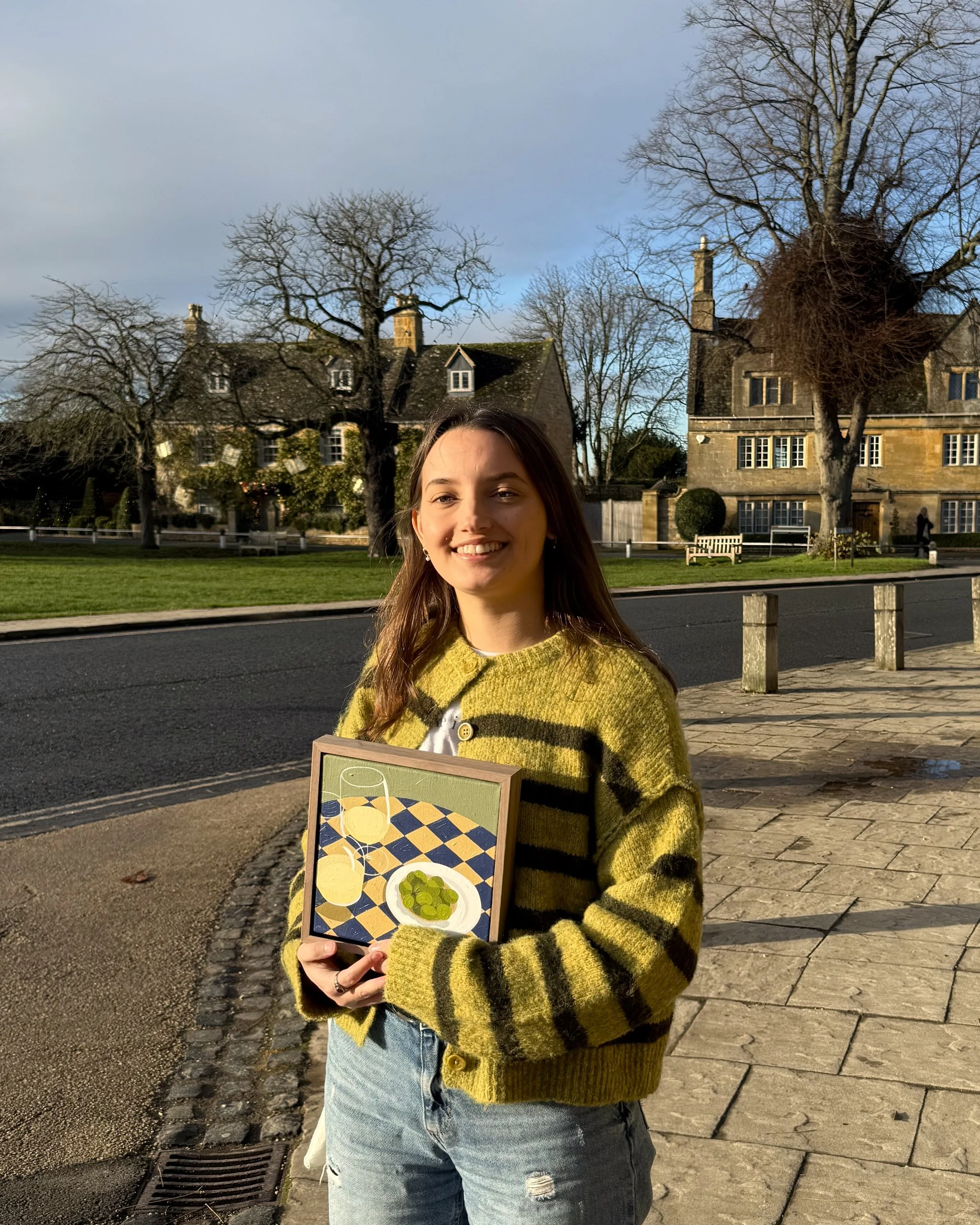 A young woman with long brown hair smiling and holding a framed still life painting. She is standing on a sidewalk in front of Cotswold stone buildings with bare trees during daytime.