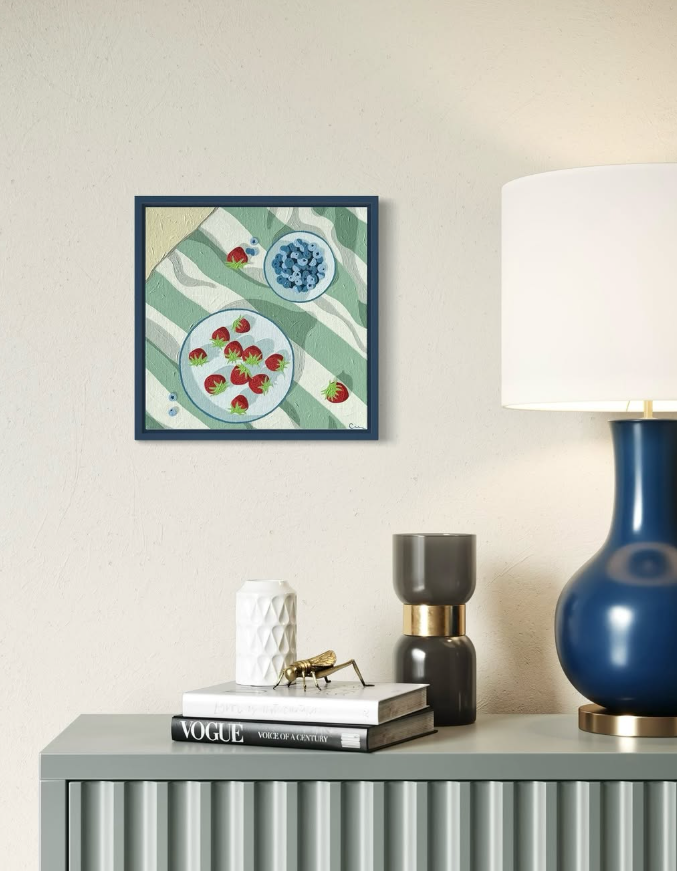 A framed artwork of strawberries and blueberries on a striped green and white background hangs above a modern sideboard. On the sideboard, there is a white textured vase, a gold-colored insect sculpture, a black and gold vase, and a large blue lamp with a white shade.